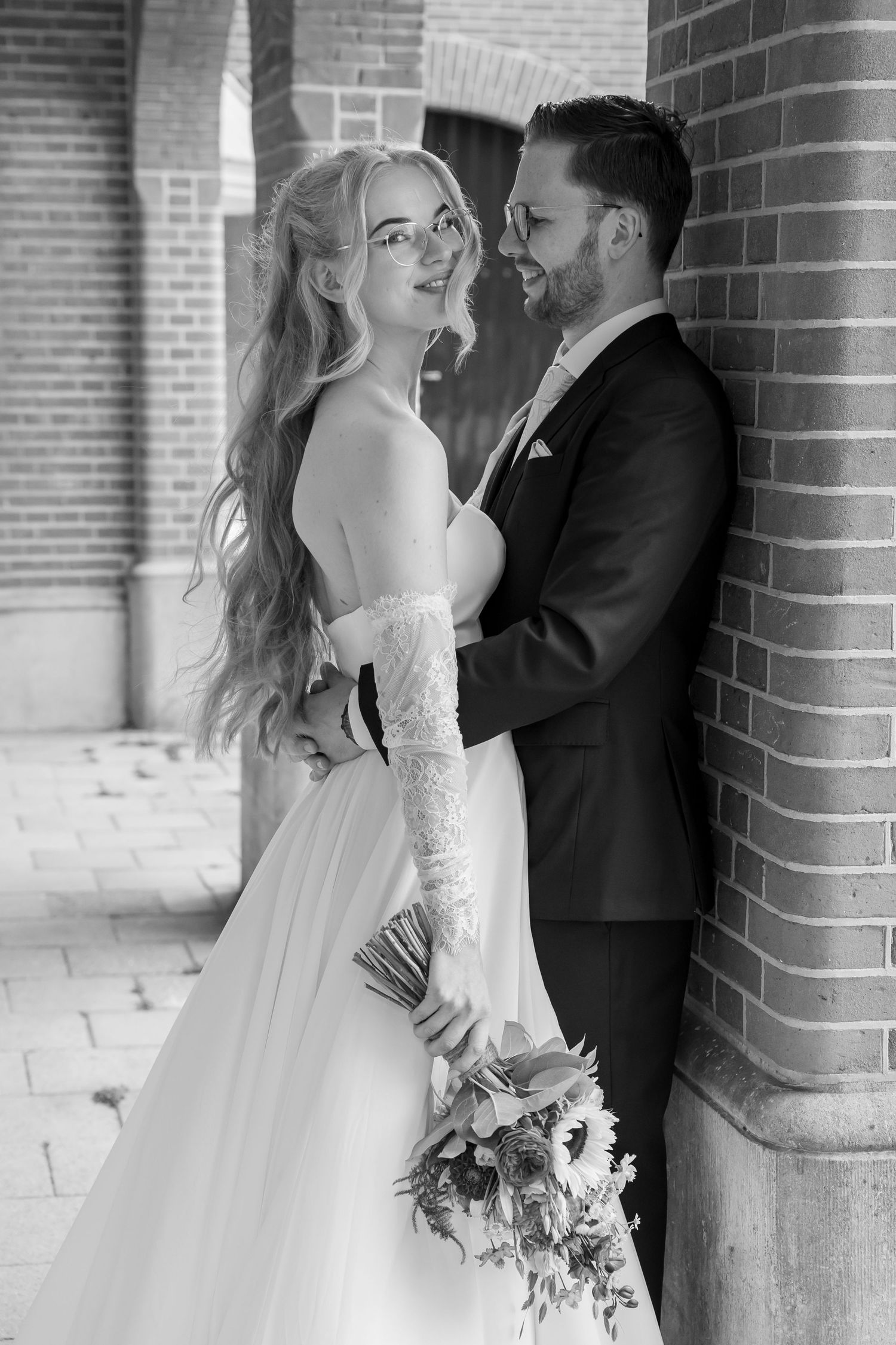 Black and white photo of bride holding bouquet smiling at groom leaning against brick column.