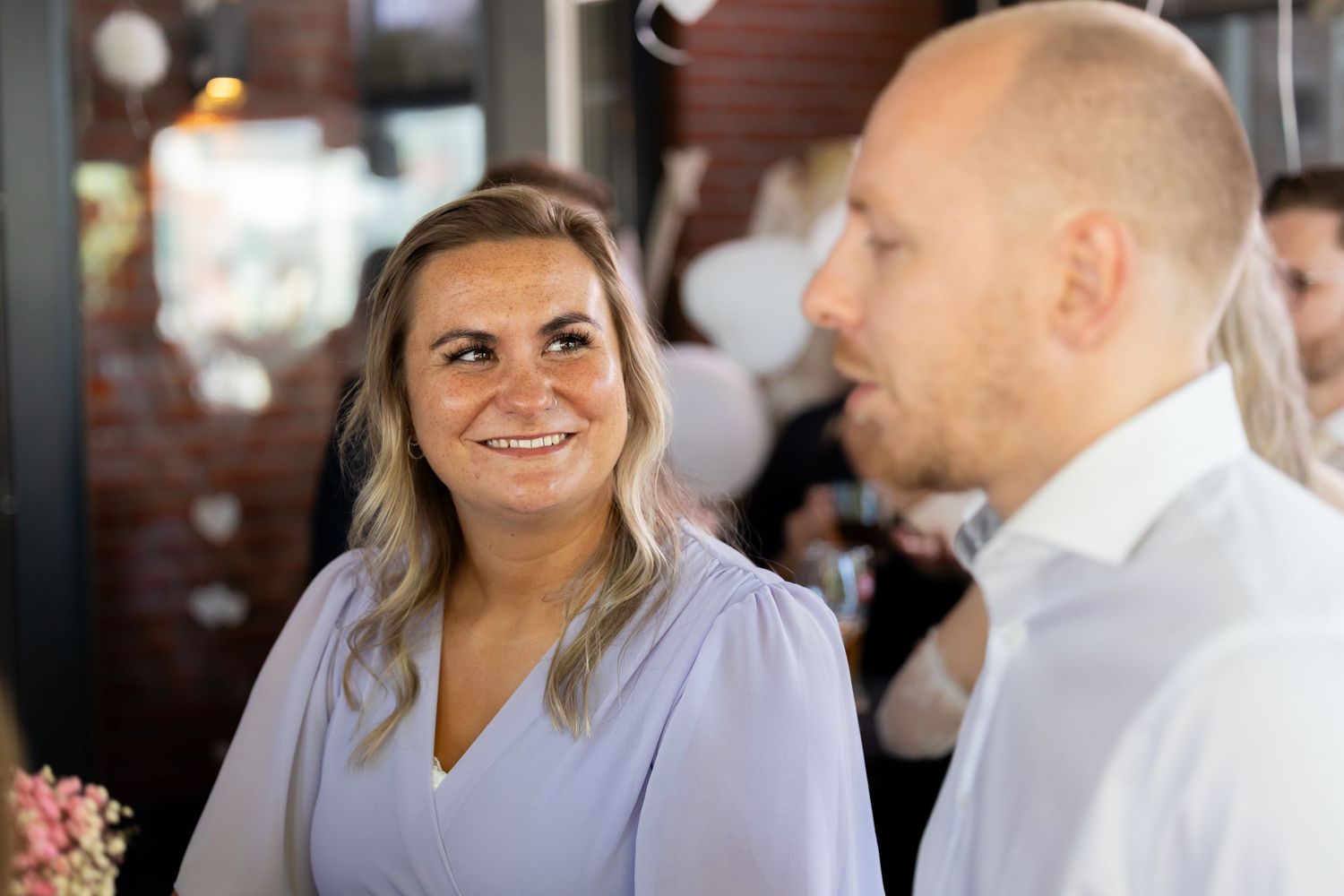 A smiling woman in a lila dress gazes at a man in a white shirt at an indoor event.