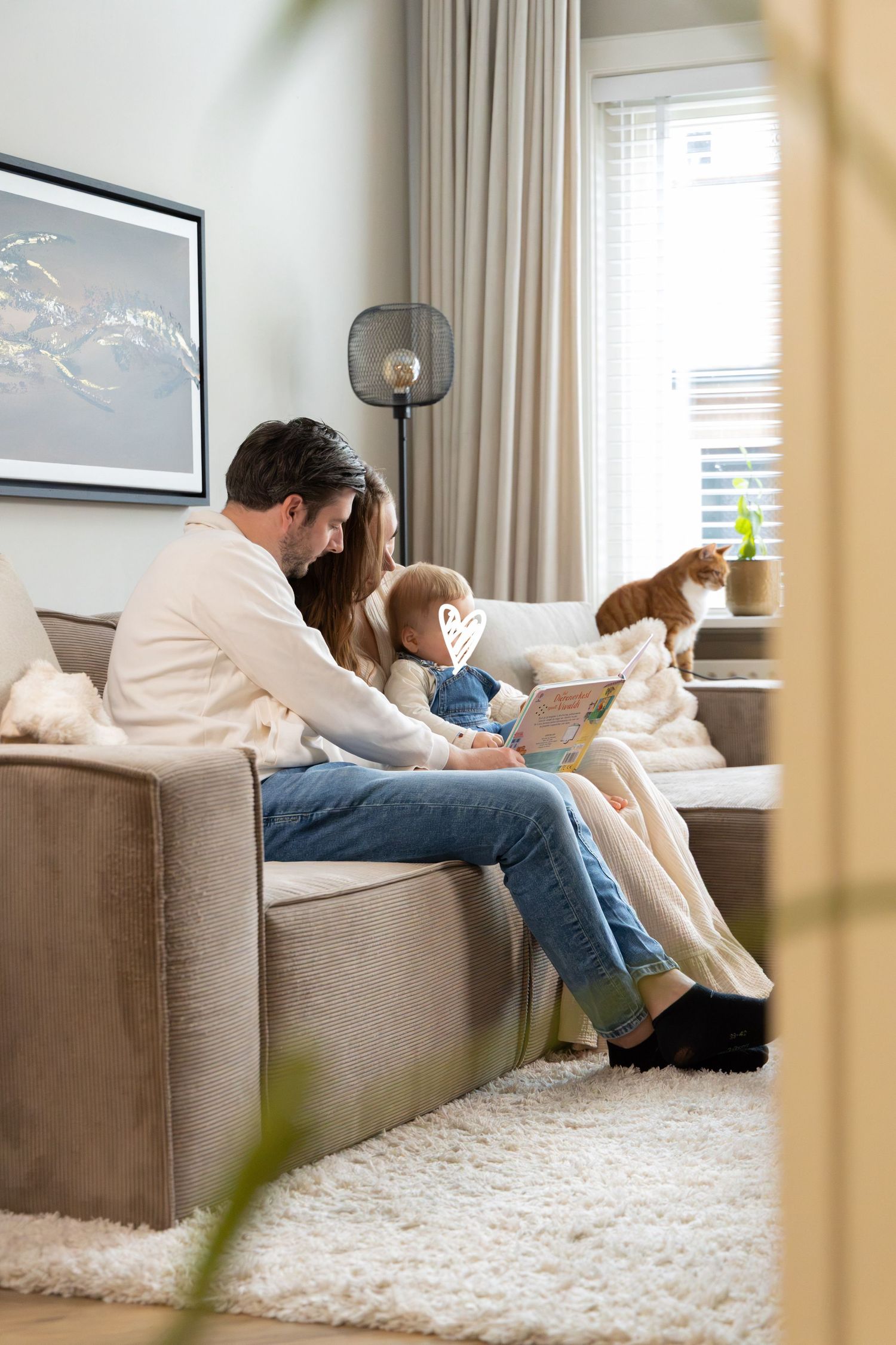 Family sitting on a cozy beige sofa reading a book together in a modern living room with cats nearby.