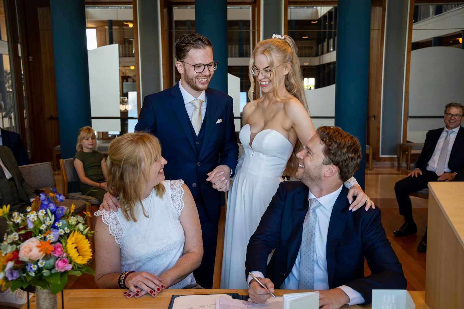 Couple in wedding attire smiling at guests signing documents during an intimate civil ceremony indoors.