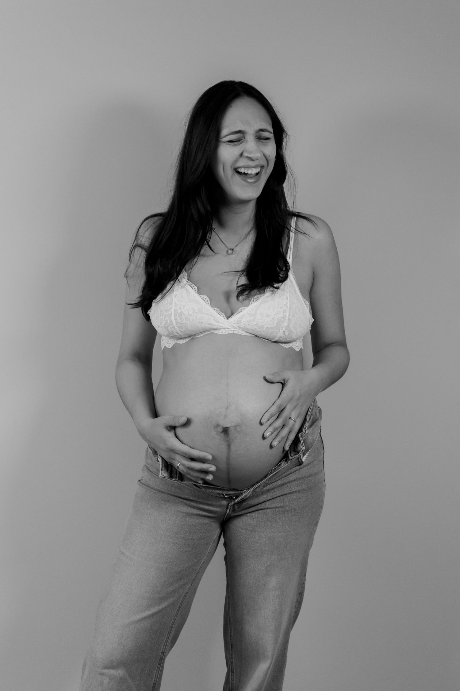 Black and white maternity portrait of a laughing pregnant woman with long dark hair cradling her bare belly in a lace bralette and jeans.