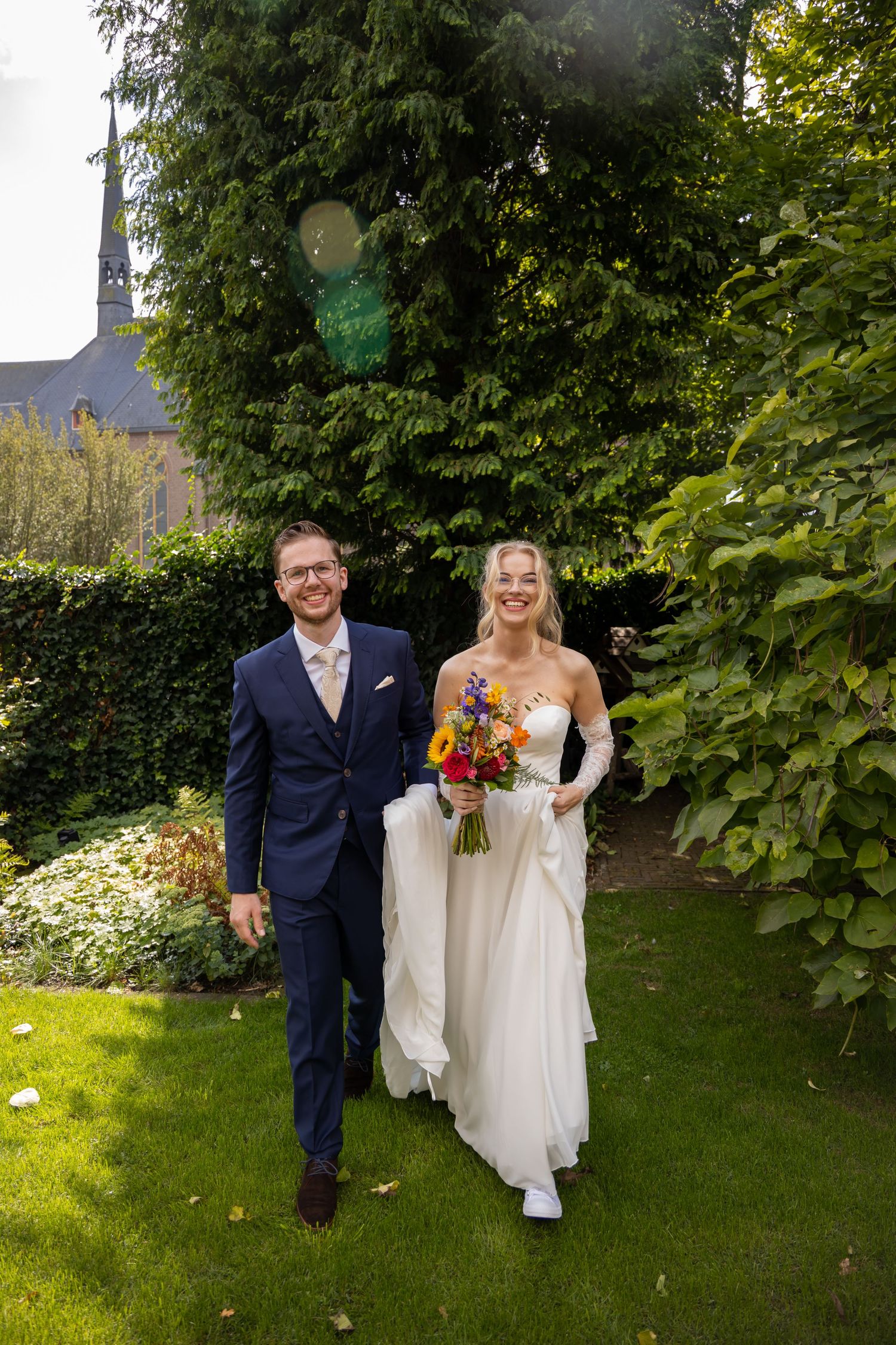 Smiling bride and groom walking through a lush garden, bride holding a colorful bouquet in a white gown.
