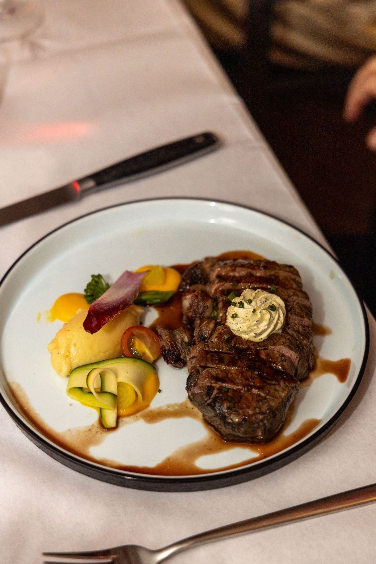 Grilled steak topped with herb butter, served with colorful vegetables and rich sauce on a white plate.