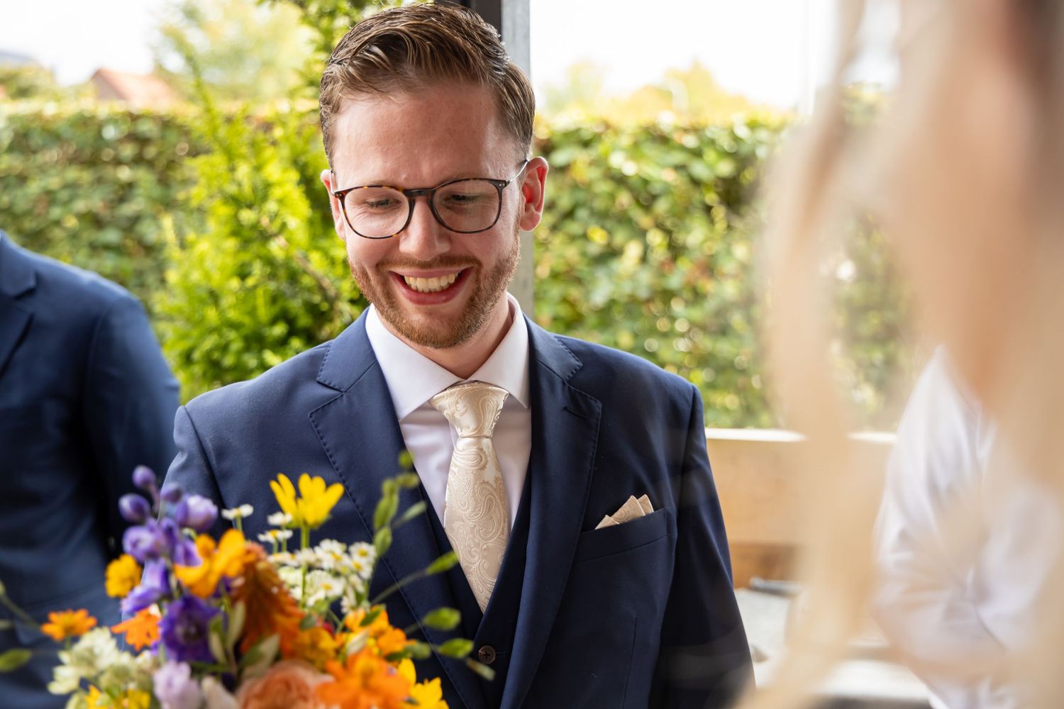 Smiling groom in navy suit and glasses holding colorful wedding bouquet during first look.