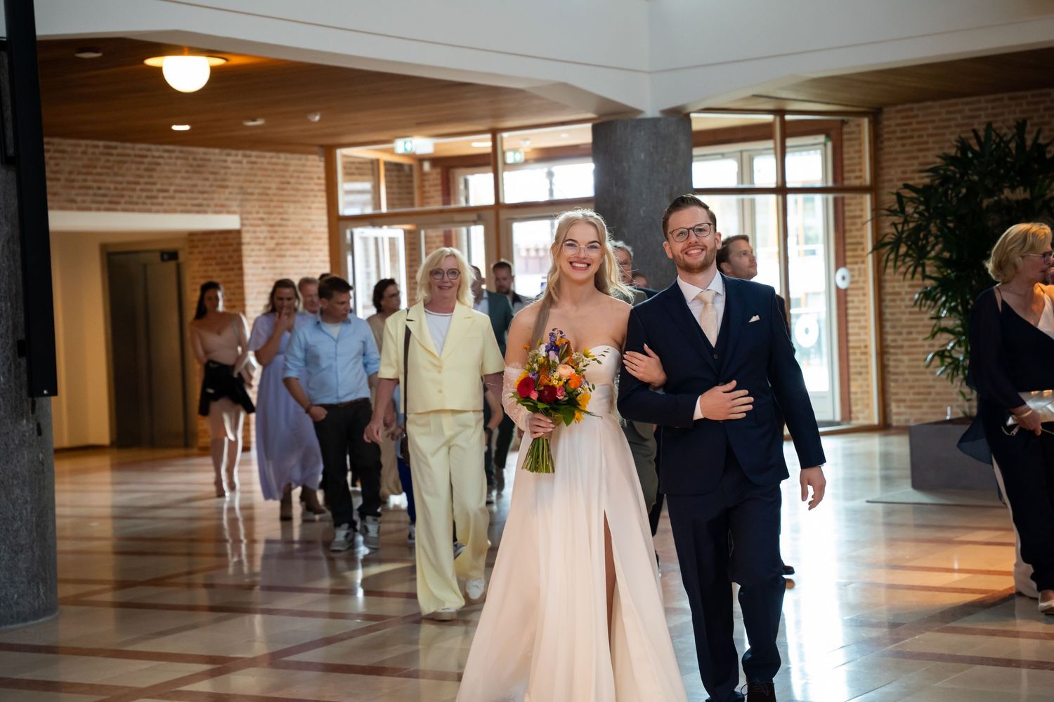 Bride in white gown holding bouquet walks with groom in navy suit through elegant venue lobby before wedding ceremony.