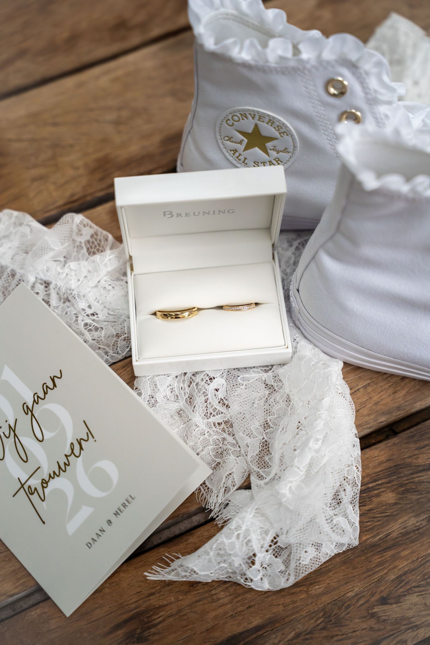 Gold wedding ring in white Breuning box with white Converse sneakers, lace fabric, and wedding invitation on wooden table.