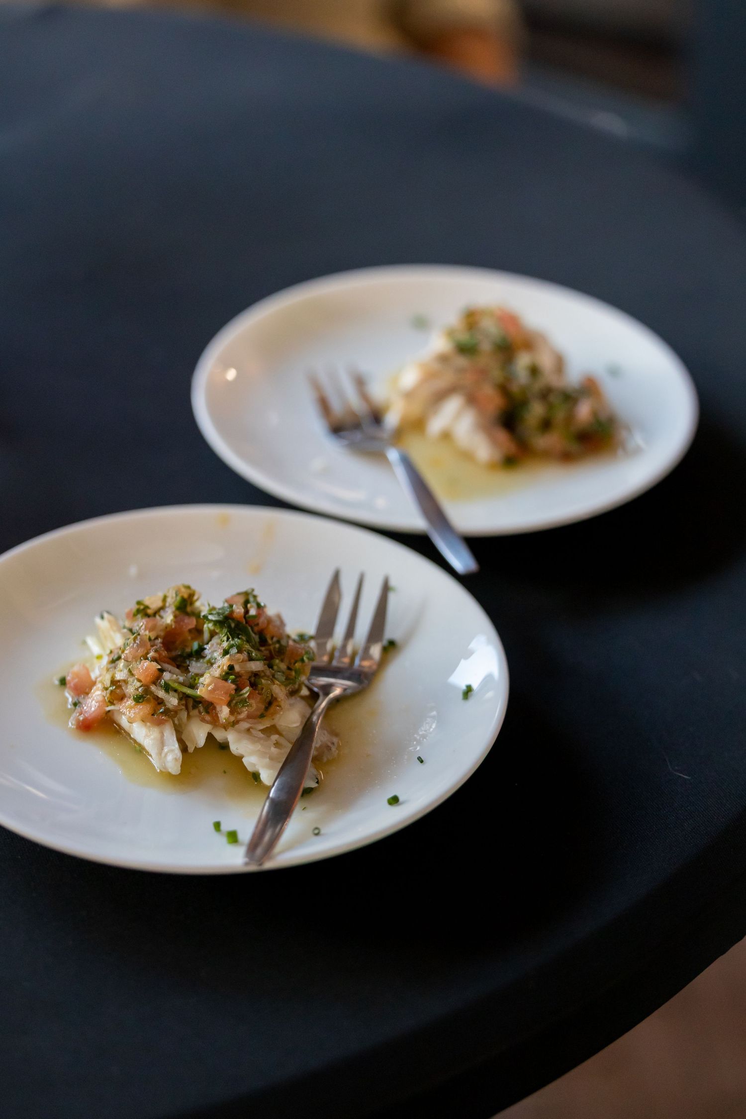 Two white plates with flaky fish topped with fresh herb salsa and forks on a dark table.