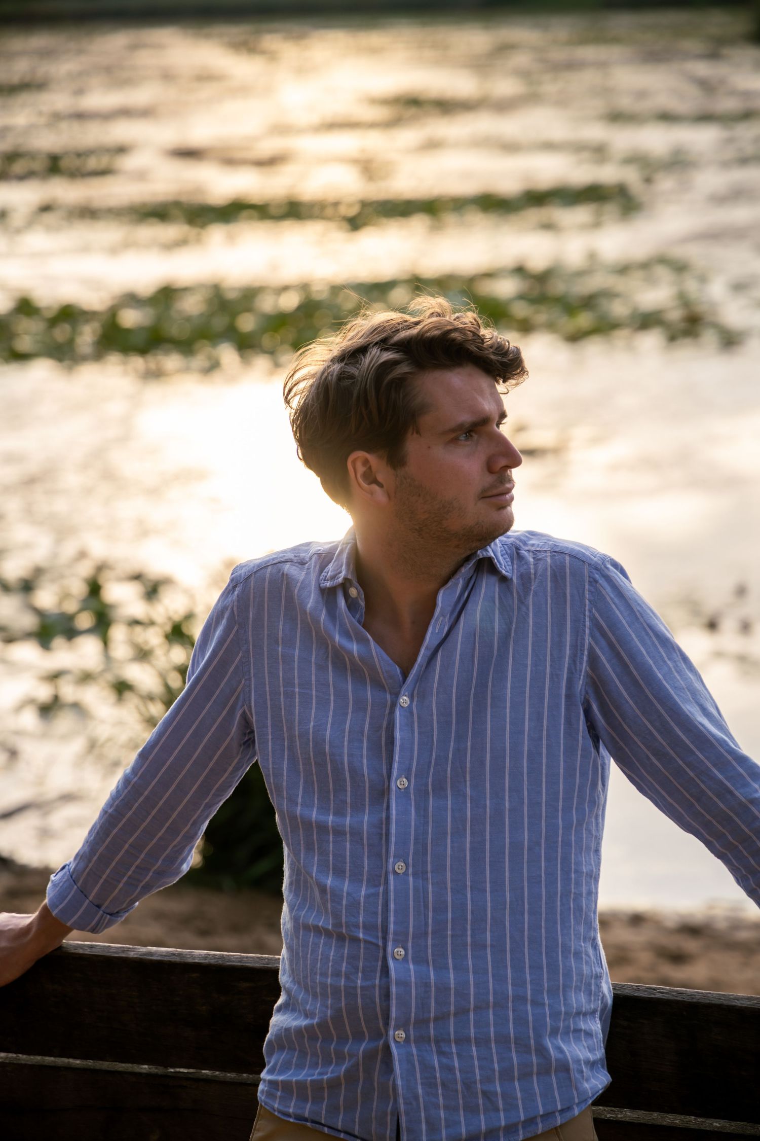 Young man in blue striped linen shirt leaning on railing by sunlit water at golden hour.