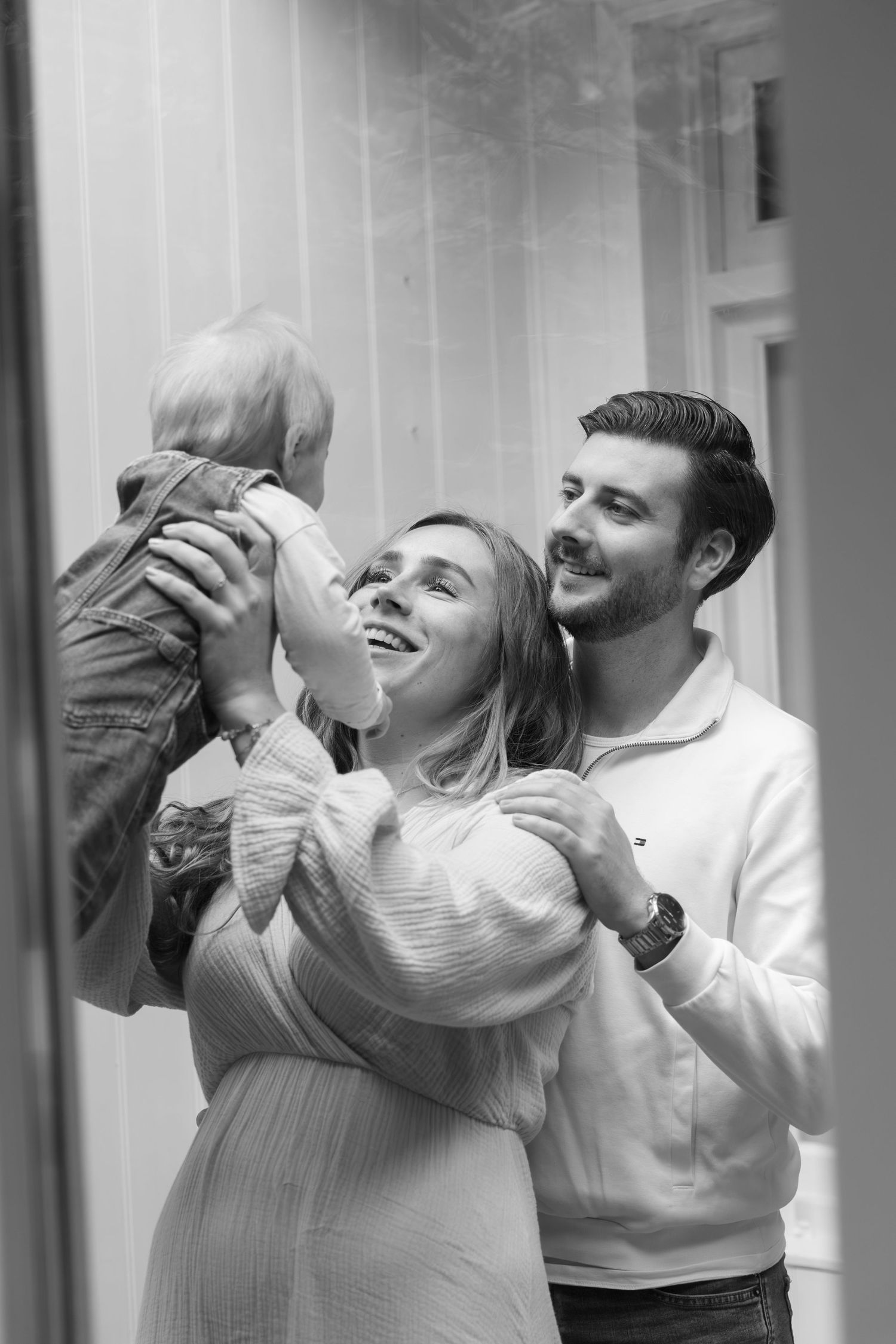 Black and white photo of happy young parents holding their baby up, smiling indoors.