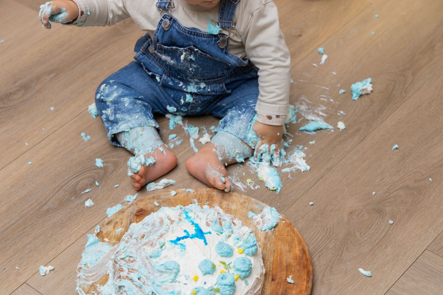 Baby in denim overalls smashing a blue and white first birthday cake on a wooden floor.