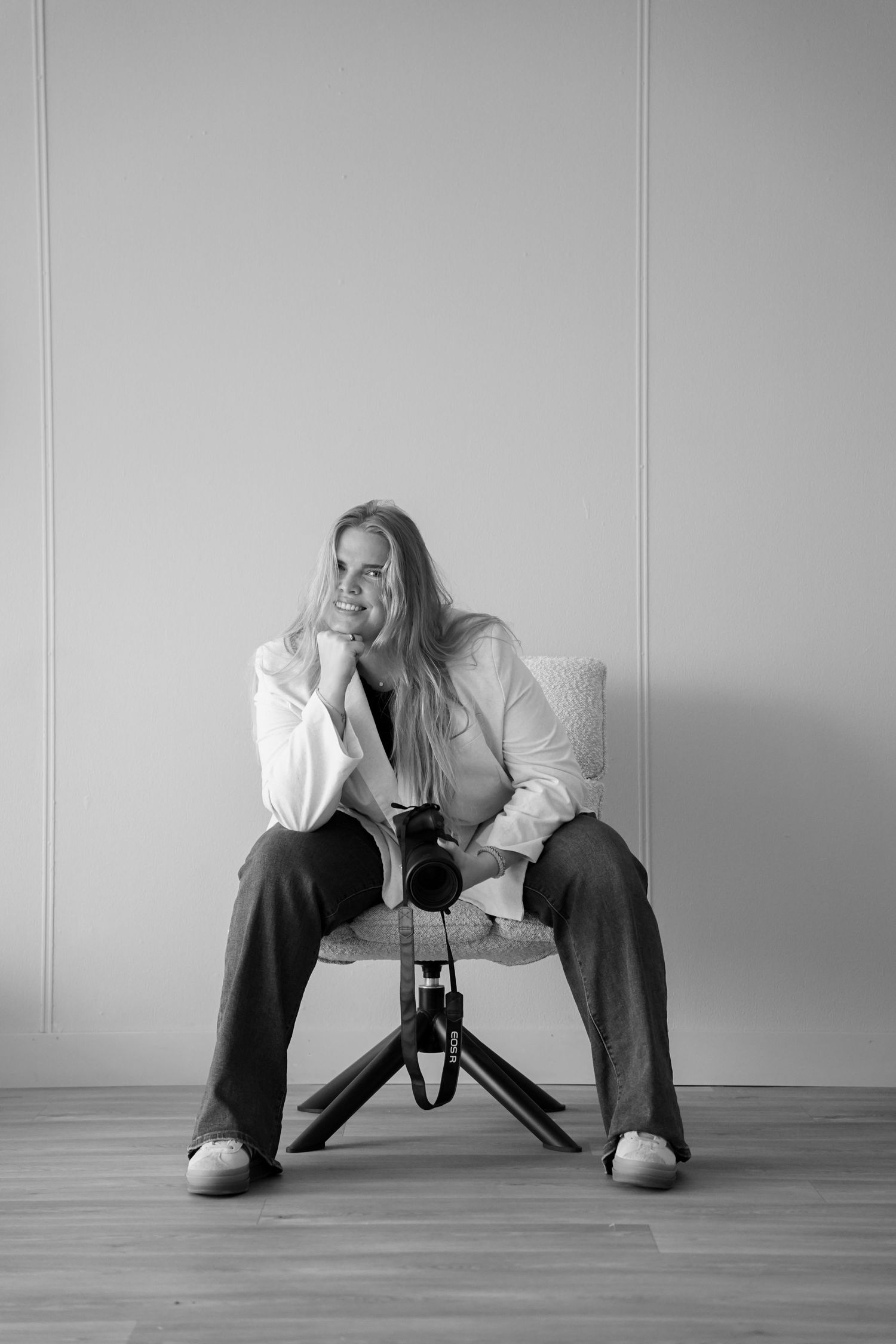 Black and white portrait of a woman sitting in a chair holding a camera, wearing a white blazer and wide-leg pants.