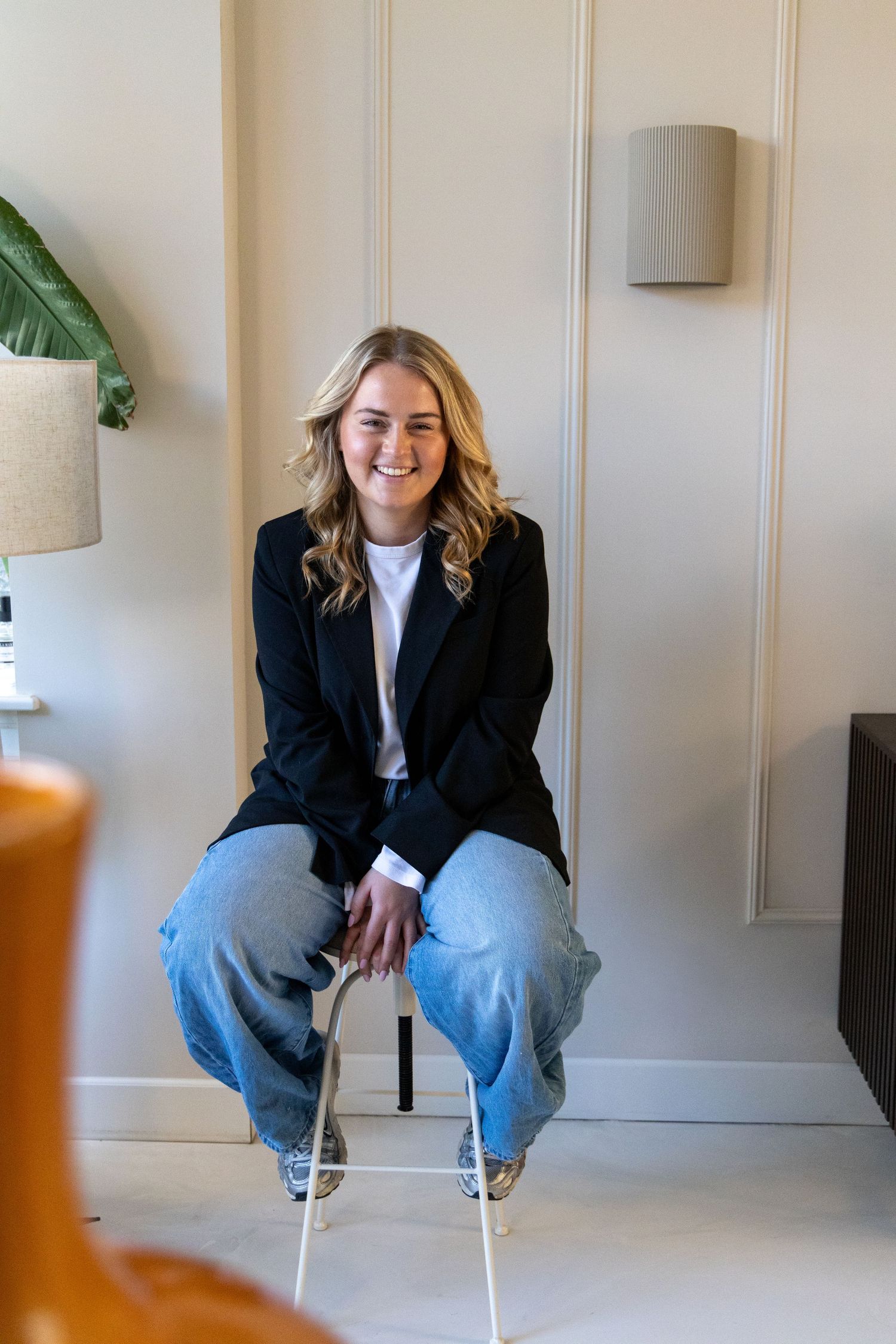 Smiling young woman with blonde hair sitting on a chair, wearing a black blazer and jeans in a bright room.
Emma - Brandingshoot