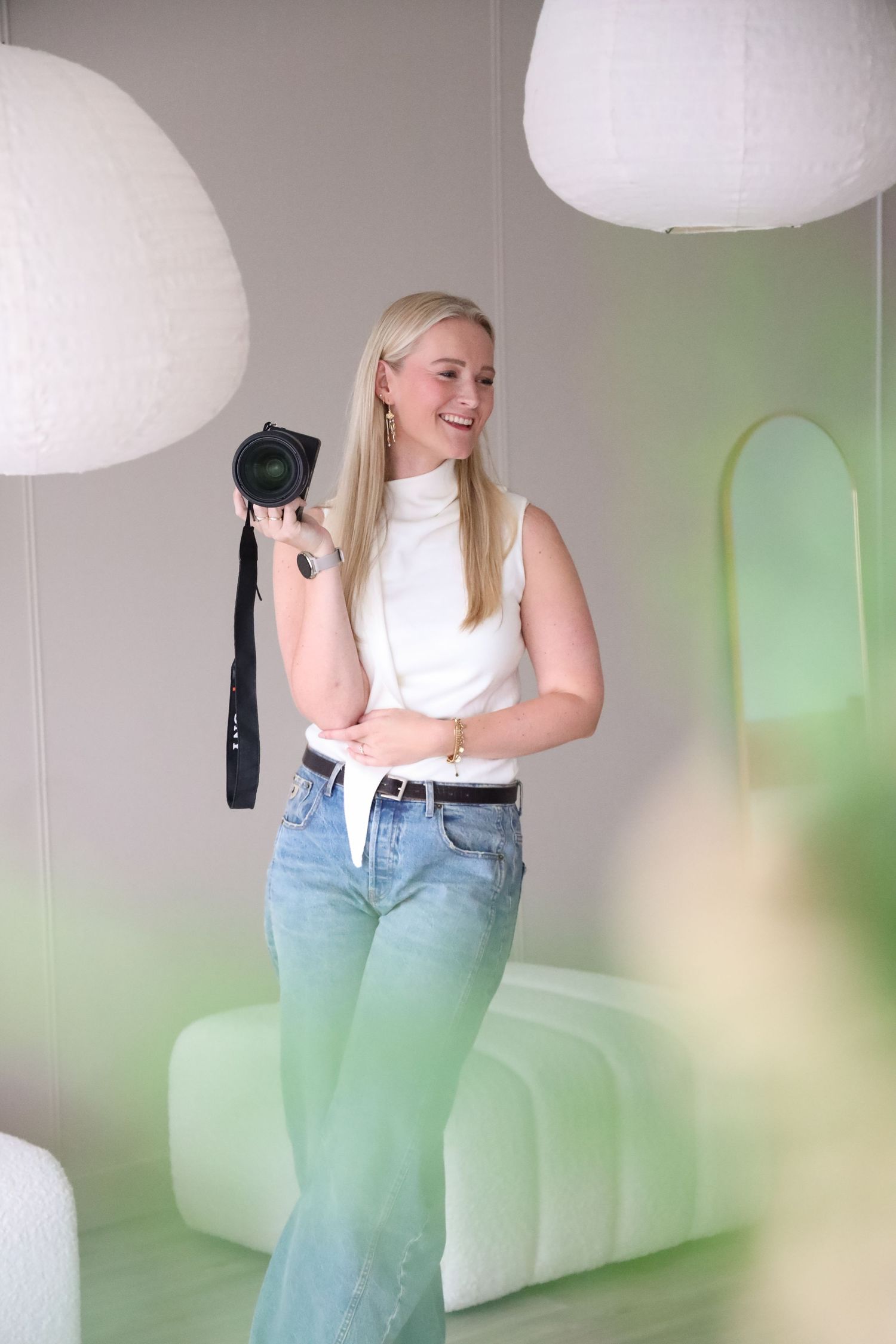 Smiling blonde woman in white top and jeans holding a DSLR camera in a modern green-toned studio space.