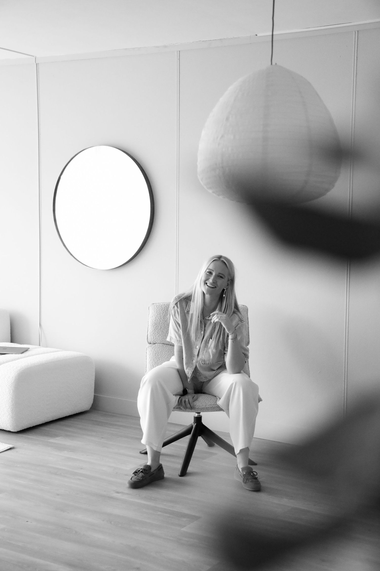 Black and white photo of a smiling blonde woman sitting in a chair in a minimalist white room with a round mirror.