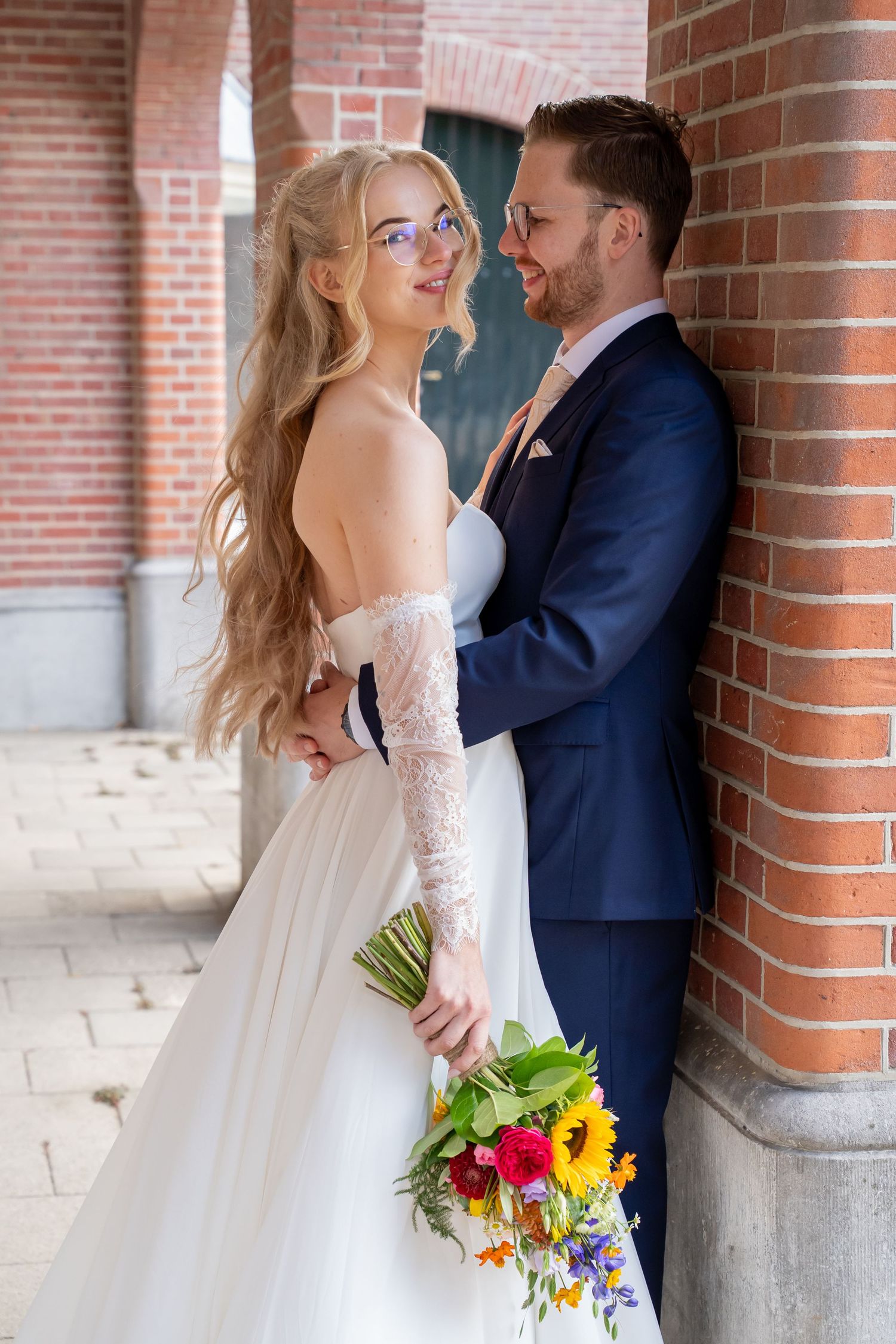 Bride in white lace gown holding colorful bouquet with groom in navy suit leaning against brick wall.