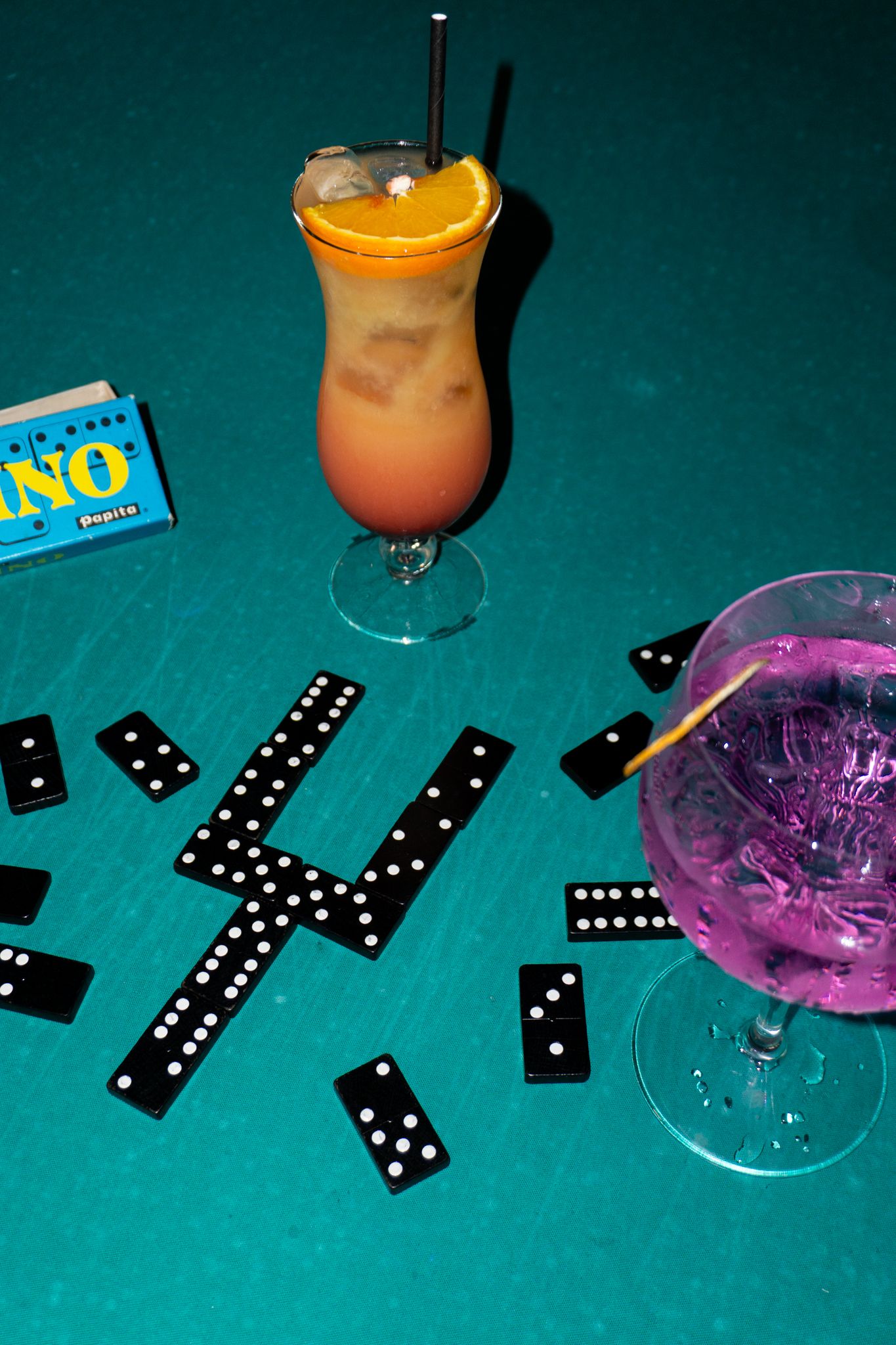Tropical cocktail with orange slice and purple drink beside dominoes game pieces on green table.
Yip's Hengelo - Brandingshoot