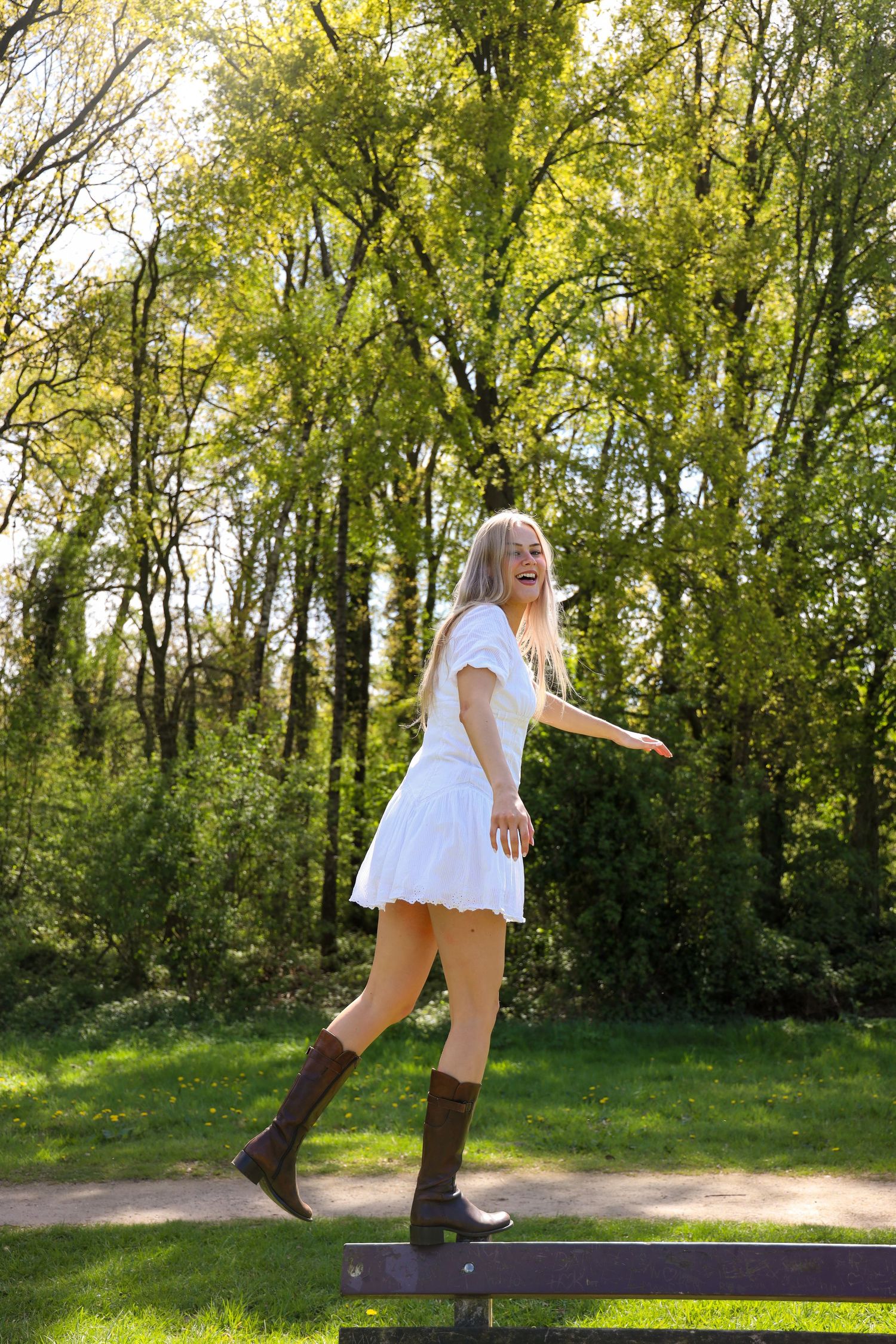 Young blonde woman in white mini dress and brown cowboy boots balancing on a bench in a sunny green park.
Romy - Soloshoot