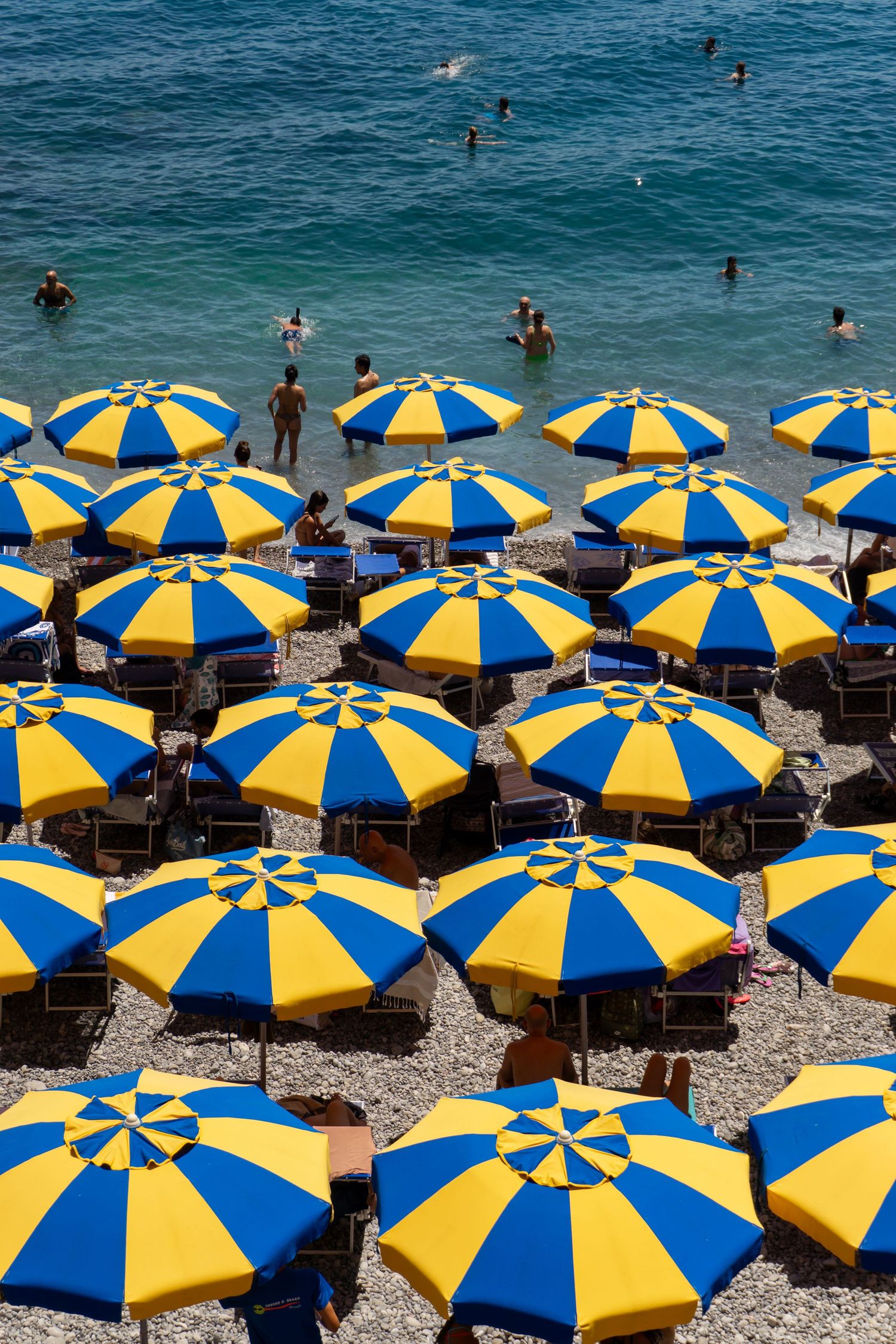 Rows of vibrant blue and yellow striped beach umbrellas on a pebble shore with swimmers in turquoise sea.
Reizen - Lynn - Amalfi