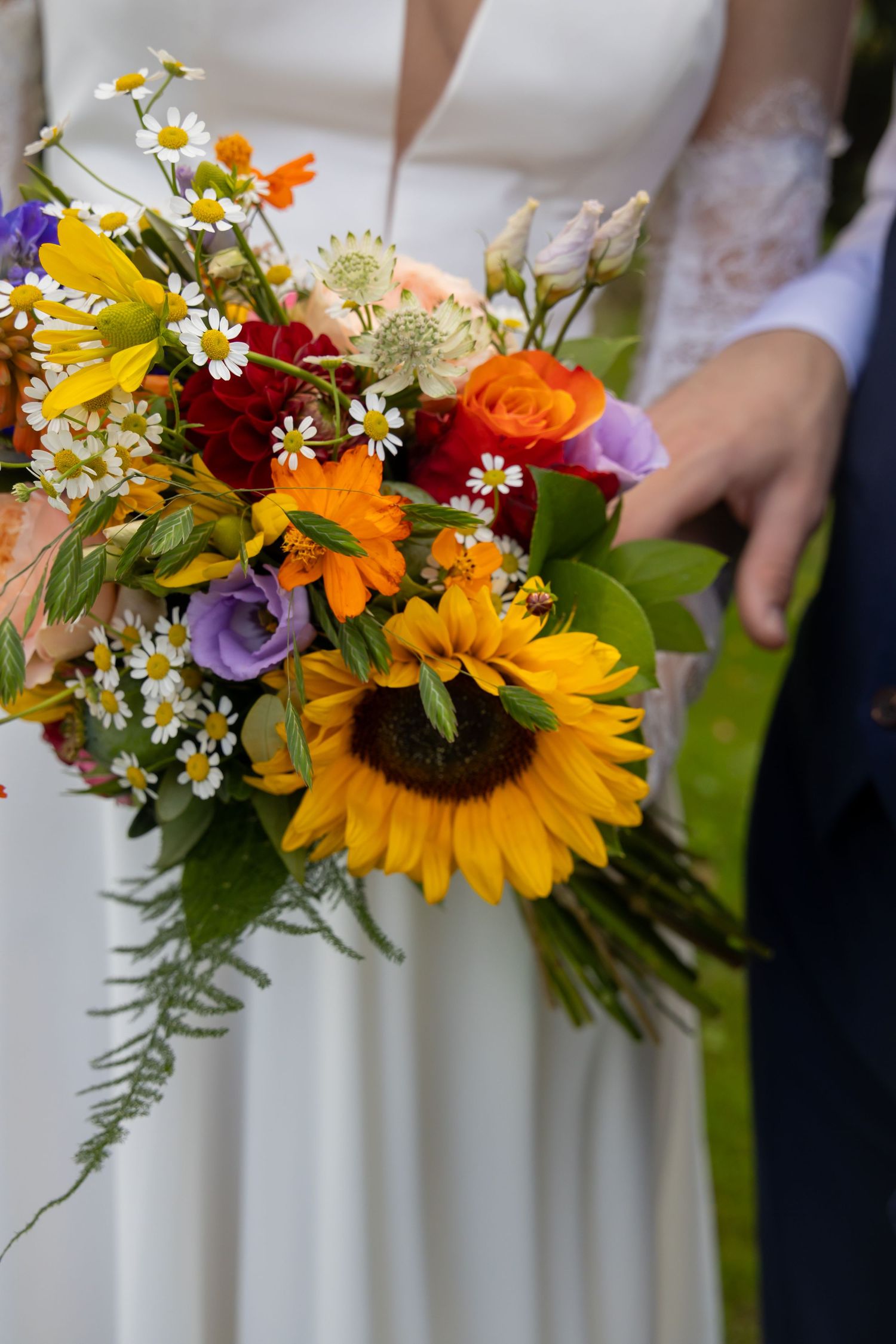 Bride holding a vibrant wedding bouquet featuring sunflowers, orange roses, daisies, and purple flowers in a colorful arrangement.