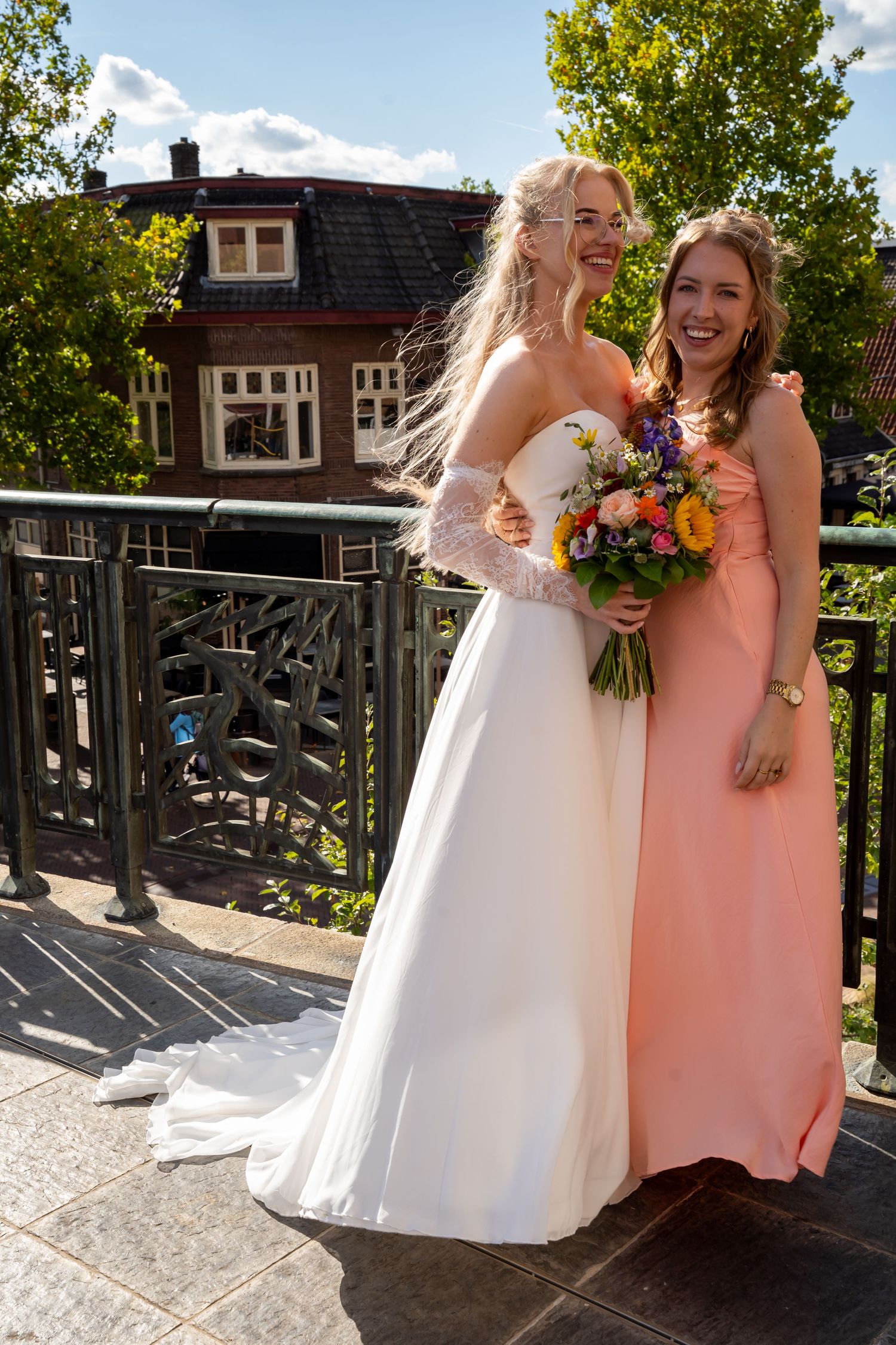 Bride in white gown and woman in peach dress smiling together, holding a colorful bouquet.
