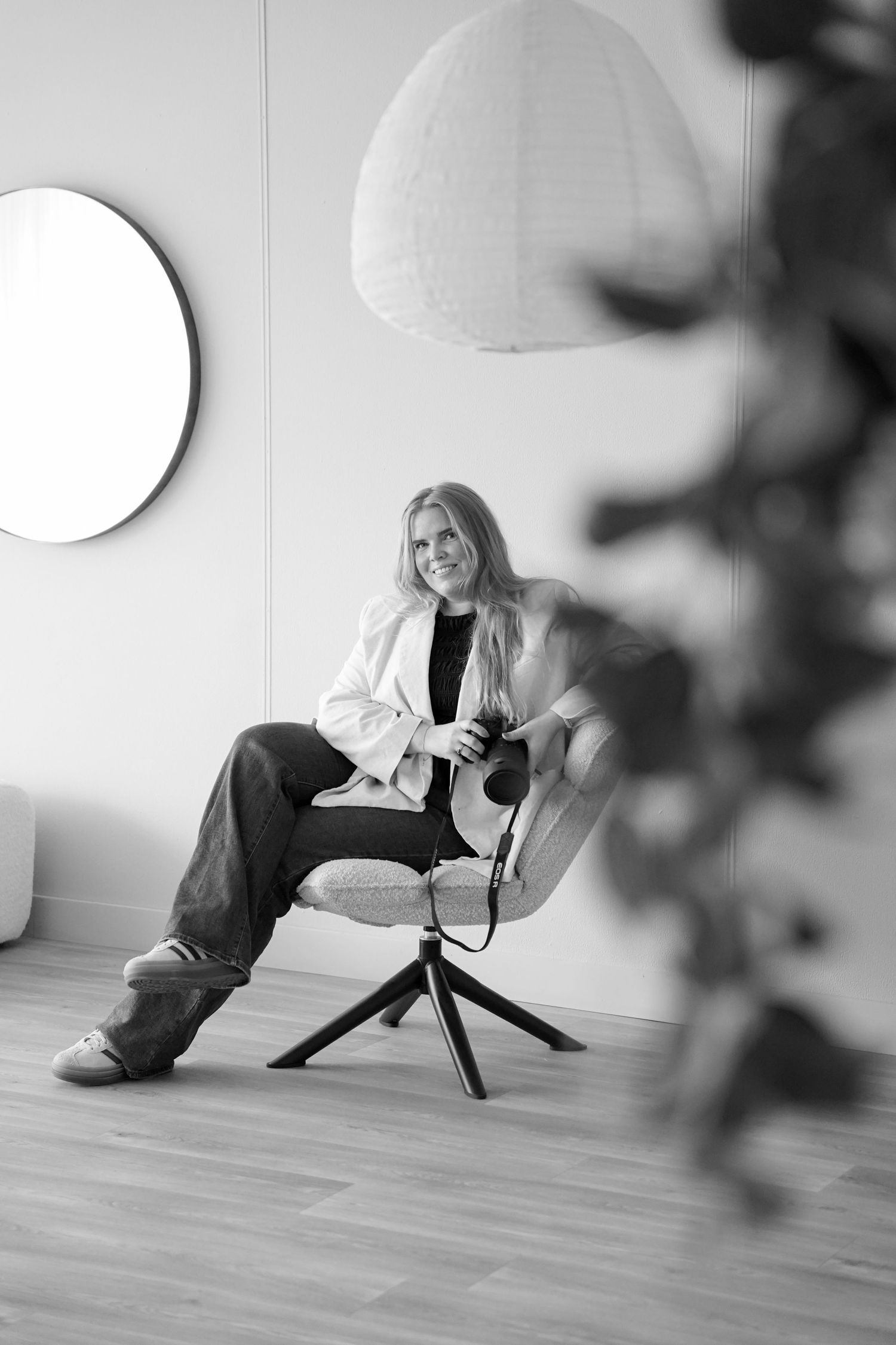Black and white photo of a smiling blonde woman sitting in a modern chair holding a camera in a minimalist room.