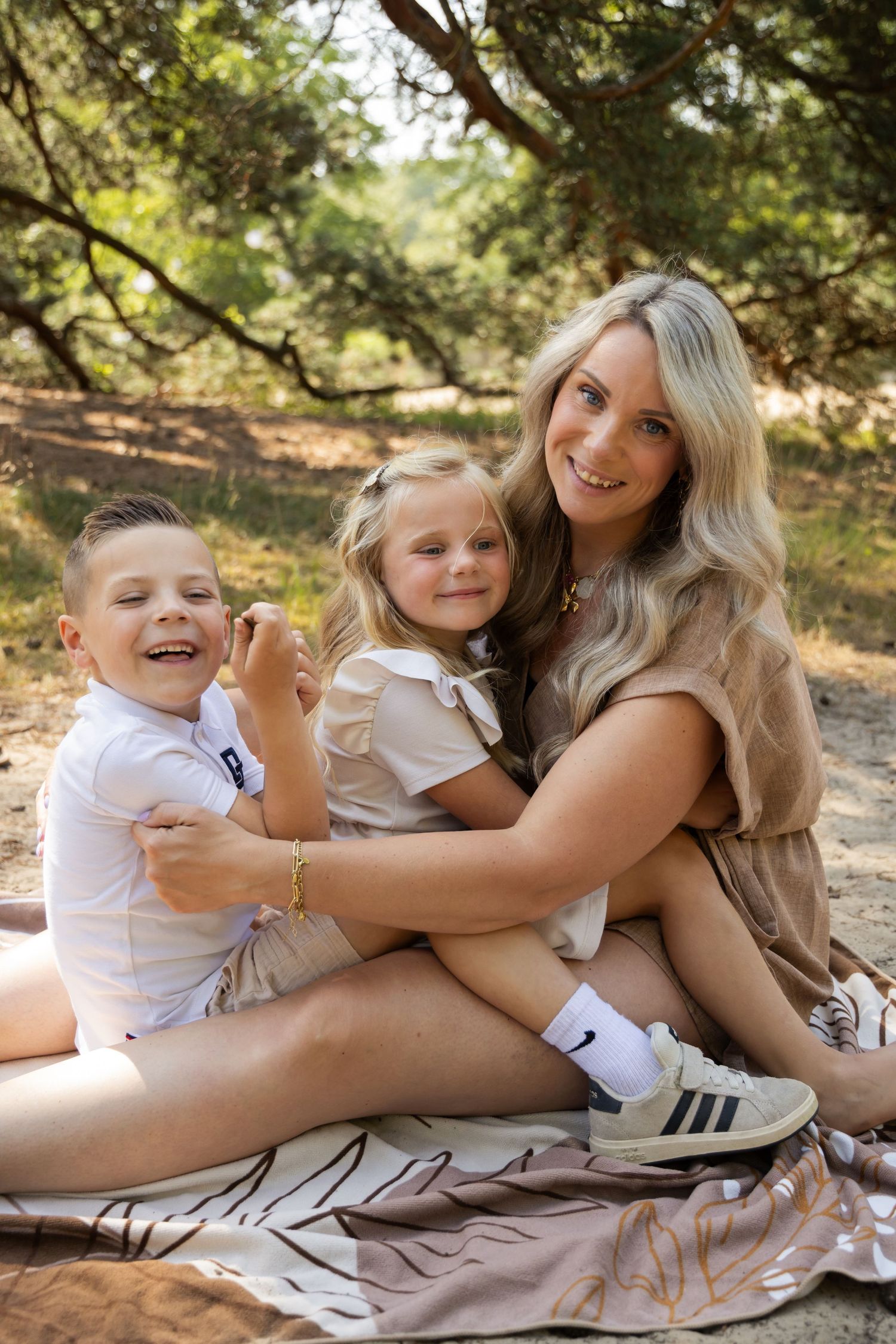 A smiling blonde woman sits outdoors on a blanket with two young children, enjoying a family picnic in a wooded area.
Familieshoot