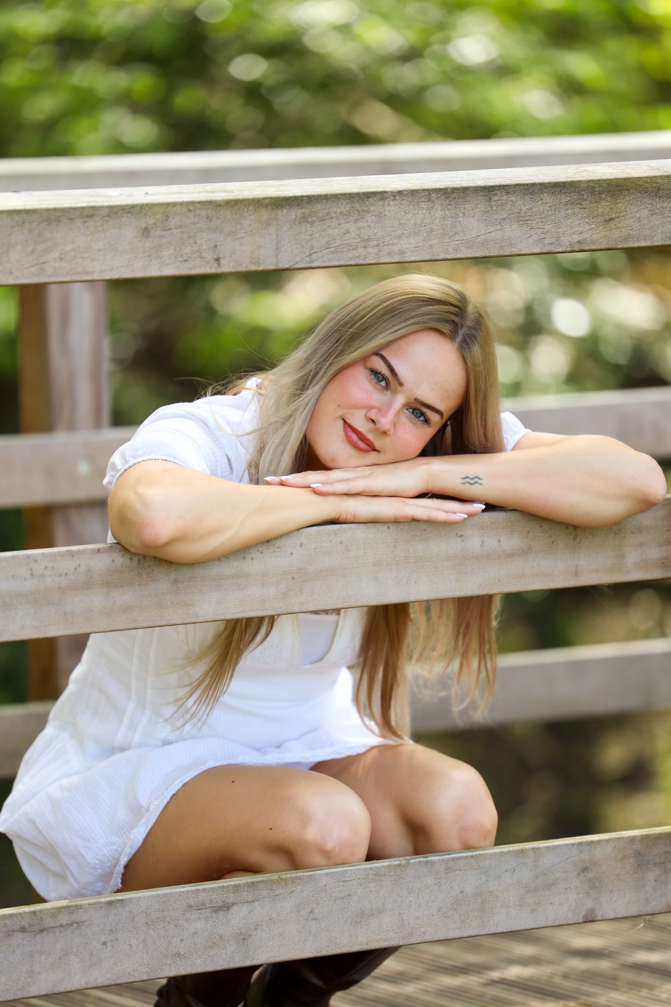 A smiling blonde woman in a white dress crouching against a wooden fence in a park.
Romy - Soloshoot