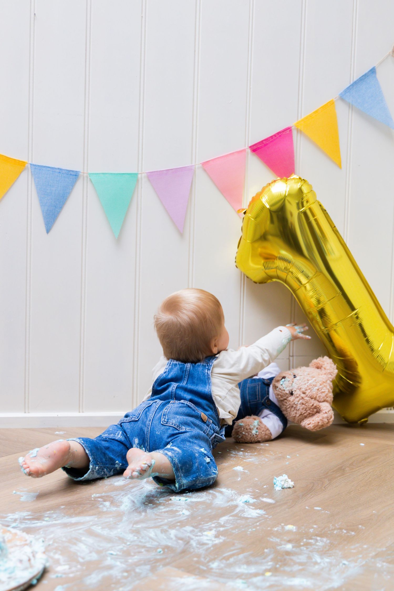 Baby in denim overalls sits on floor with teddy bear and gold number 1 balloon at first birthday party.