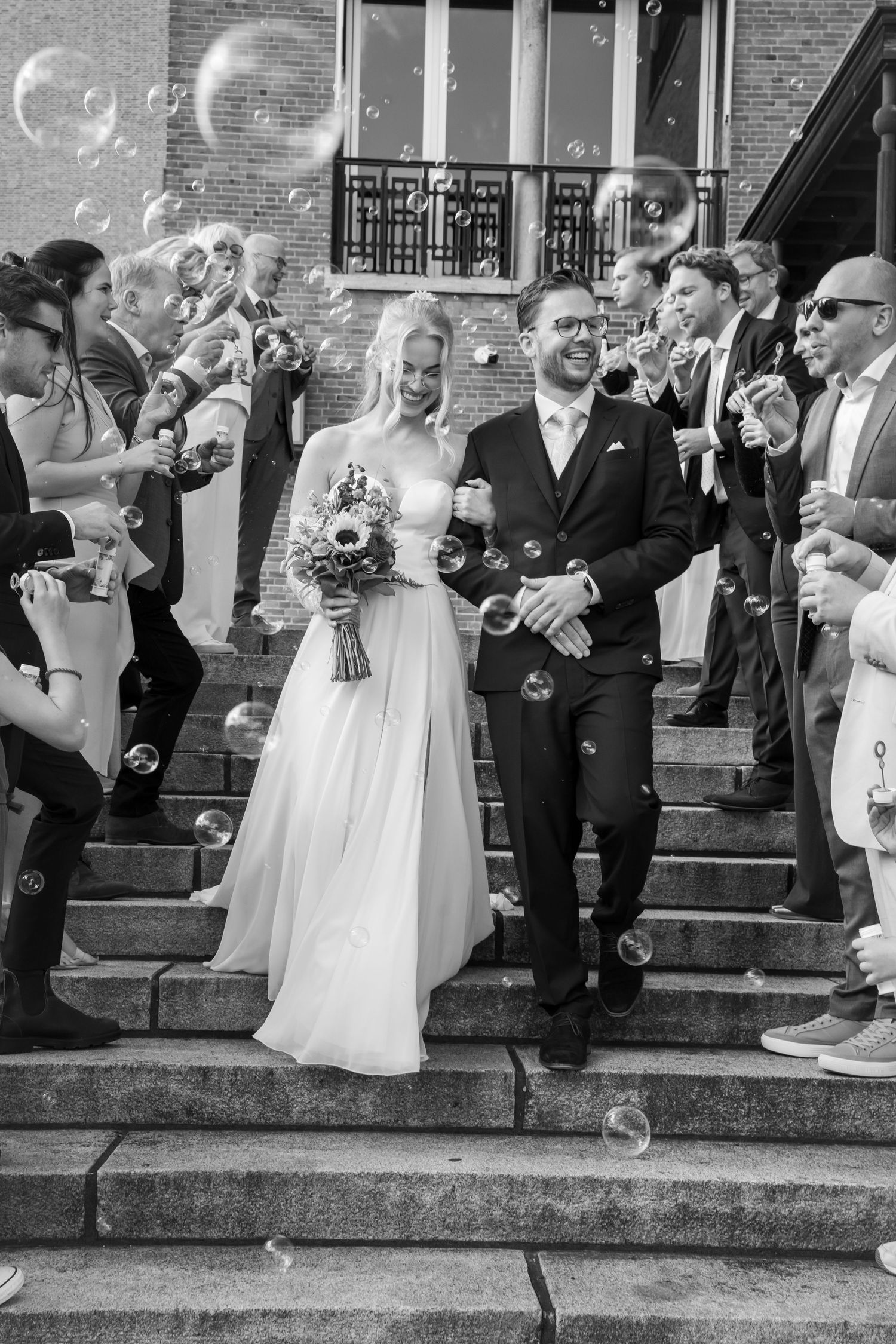Bride and groom exit steps surrounded by guests blowing bubbles in black and white photo.