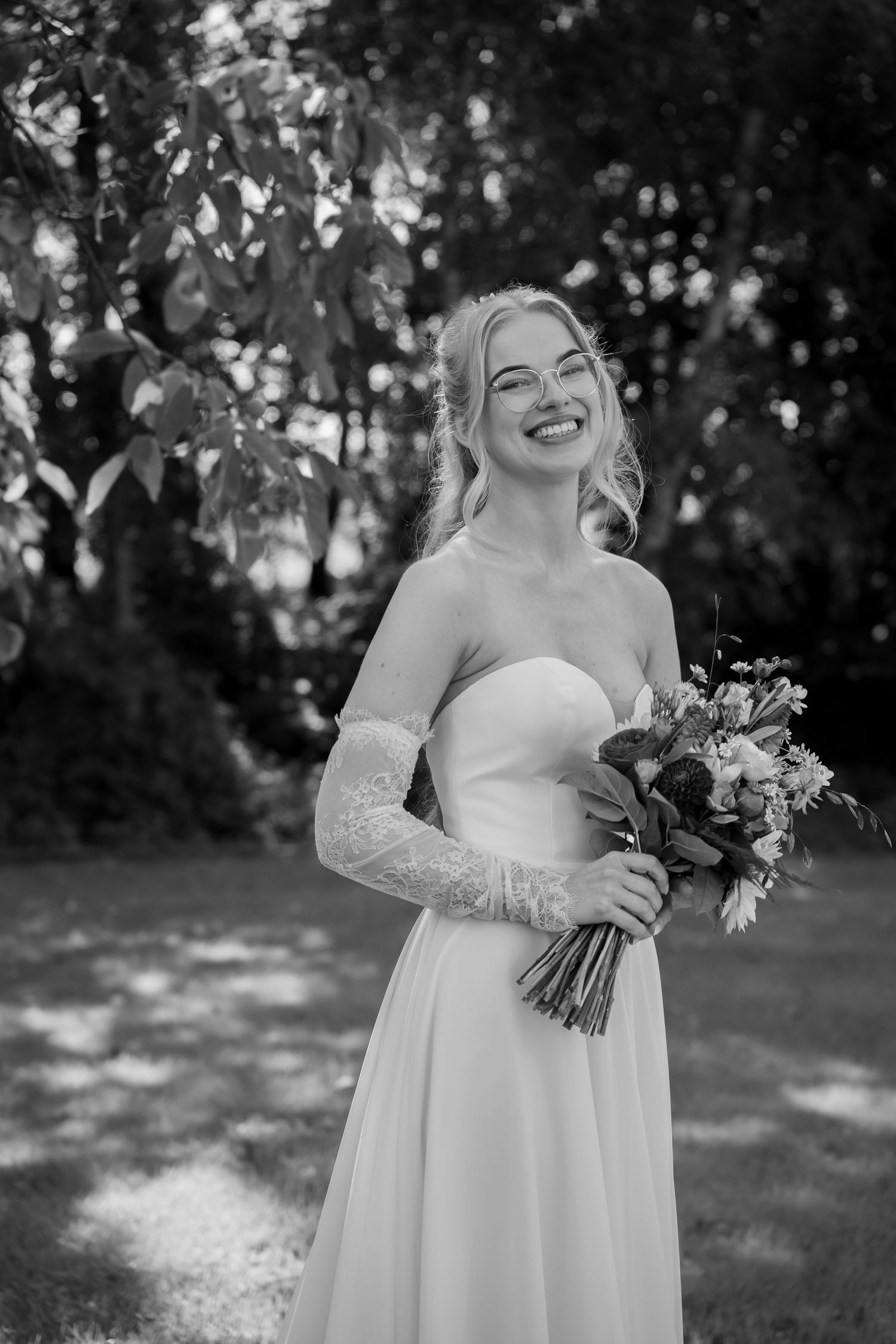 Happy bride in strapless white gown with lace gloves holds floral bouquet outdoors in black and white photo.