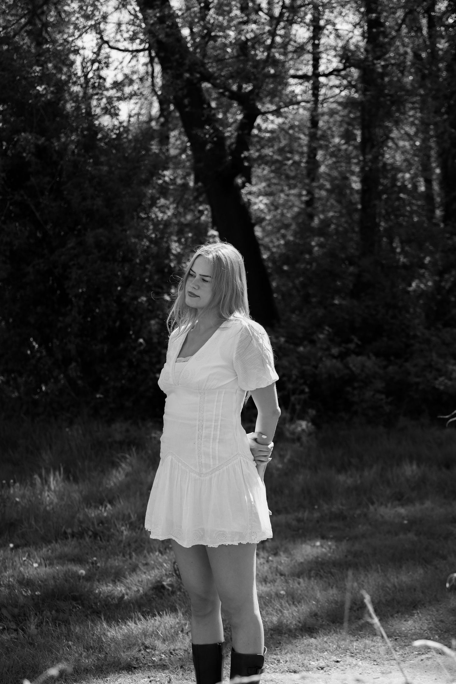 Black and white photo of a blonde woman in a white mini dress and tall boots standing in a wooded outdoor setting.
Romy - Soloshoot