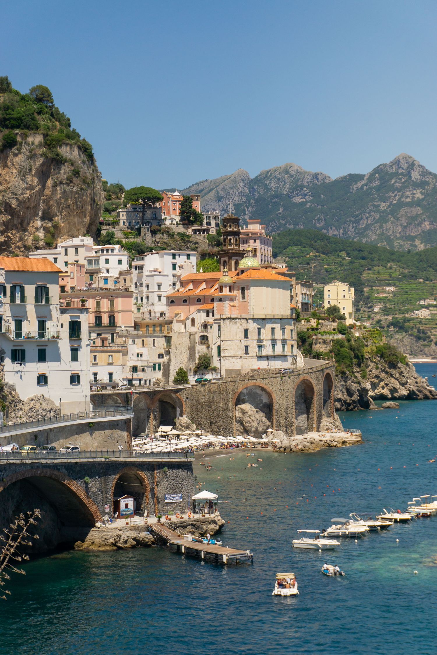 Scenic Amalfi Coast town with colorful buildings, stone bridge arches, and boats on turquoise water backed by mountains.
Lynn - Atrani