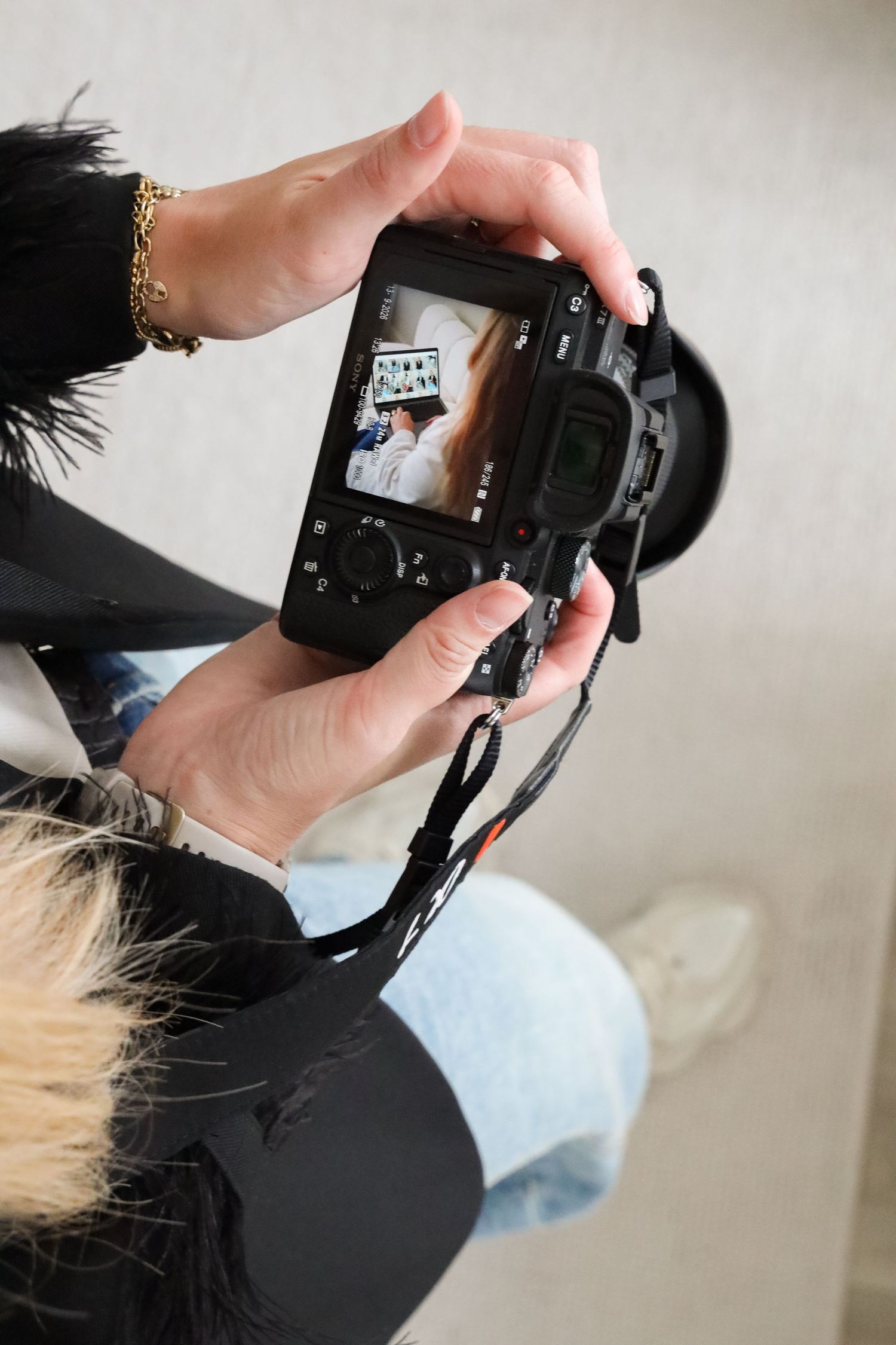 Photographer reviewing photos on DSLR camera screen while seated, wearing black jacket with fur trim and gold bracelet.