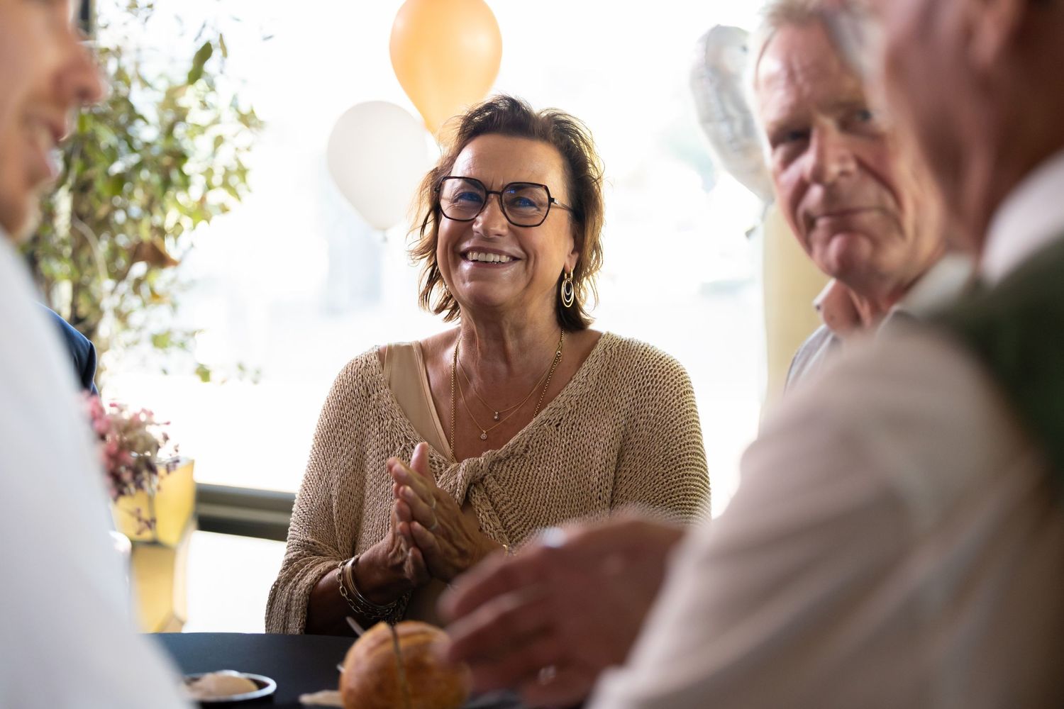 A cheerful woman with glasses claps hands at a festive celebration with balloons and friends.