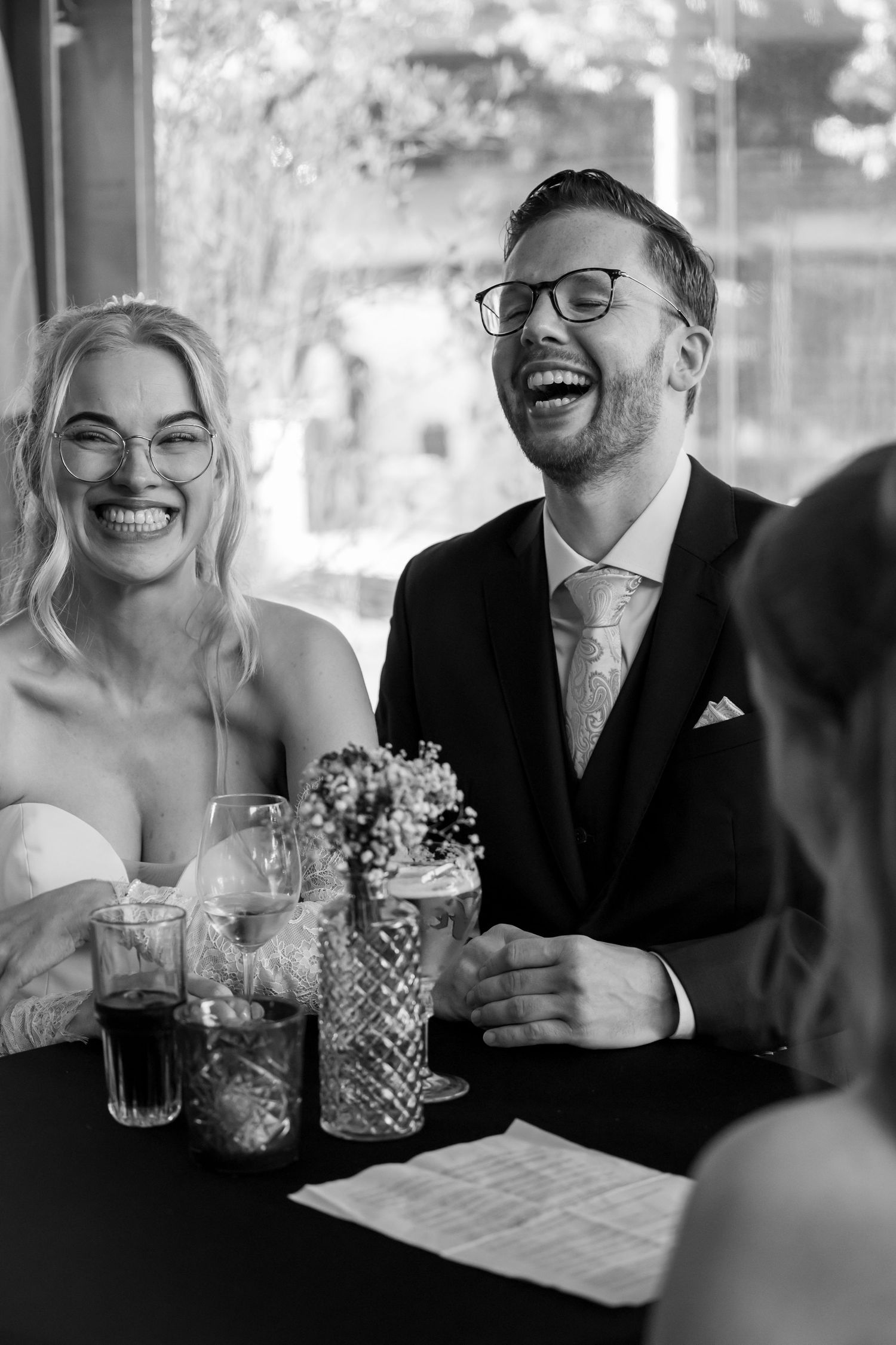 Bride and groom laughing joyfully together at reception table in black and white wedding photo.