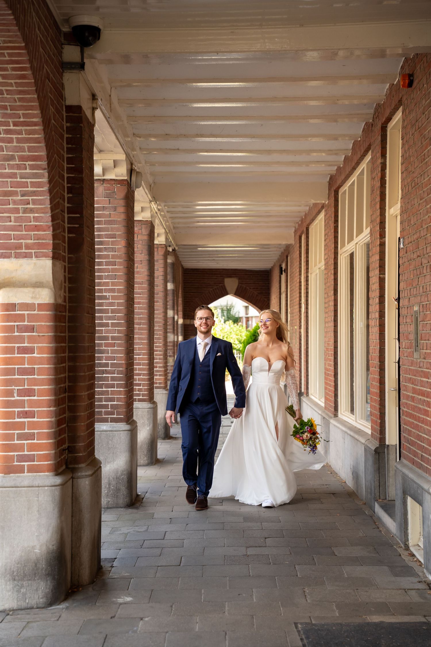 Bride and groom walking hand in hand through a brick archway corridor on their wedding day.