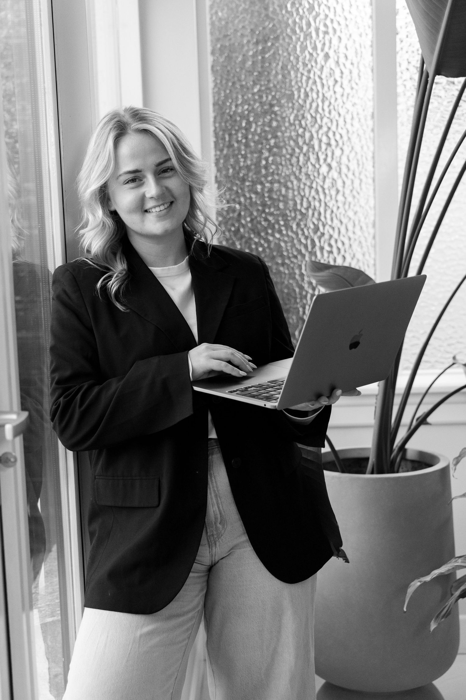 Smiling blonde woman in black blazer holding laptop, standing near window in black and white professional photo.
Emma - Brandingshoot