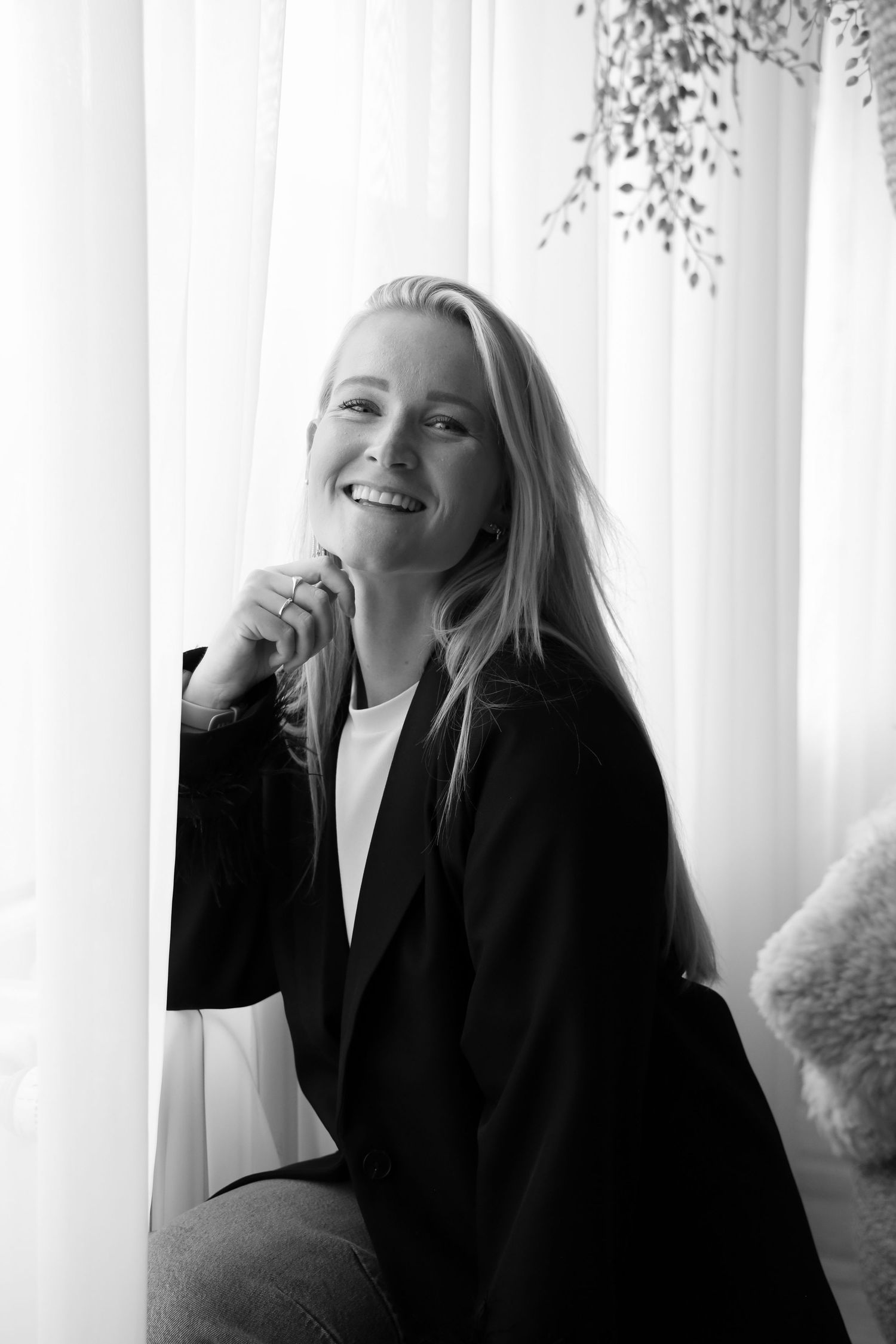 Smiling blonde woman in black blazer poses by sheer curtains in a bright, elegant black and white portrait photo.