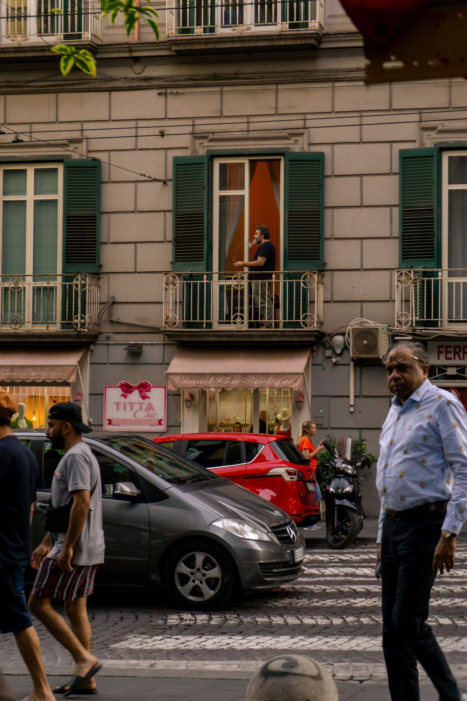 Busy Italian street scene with pedestrians, parked cars, and a man on a balcony of a classic building with green shutters.
Lynn - Napoli