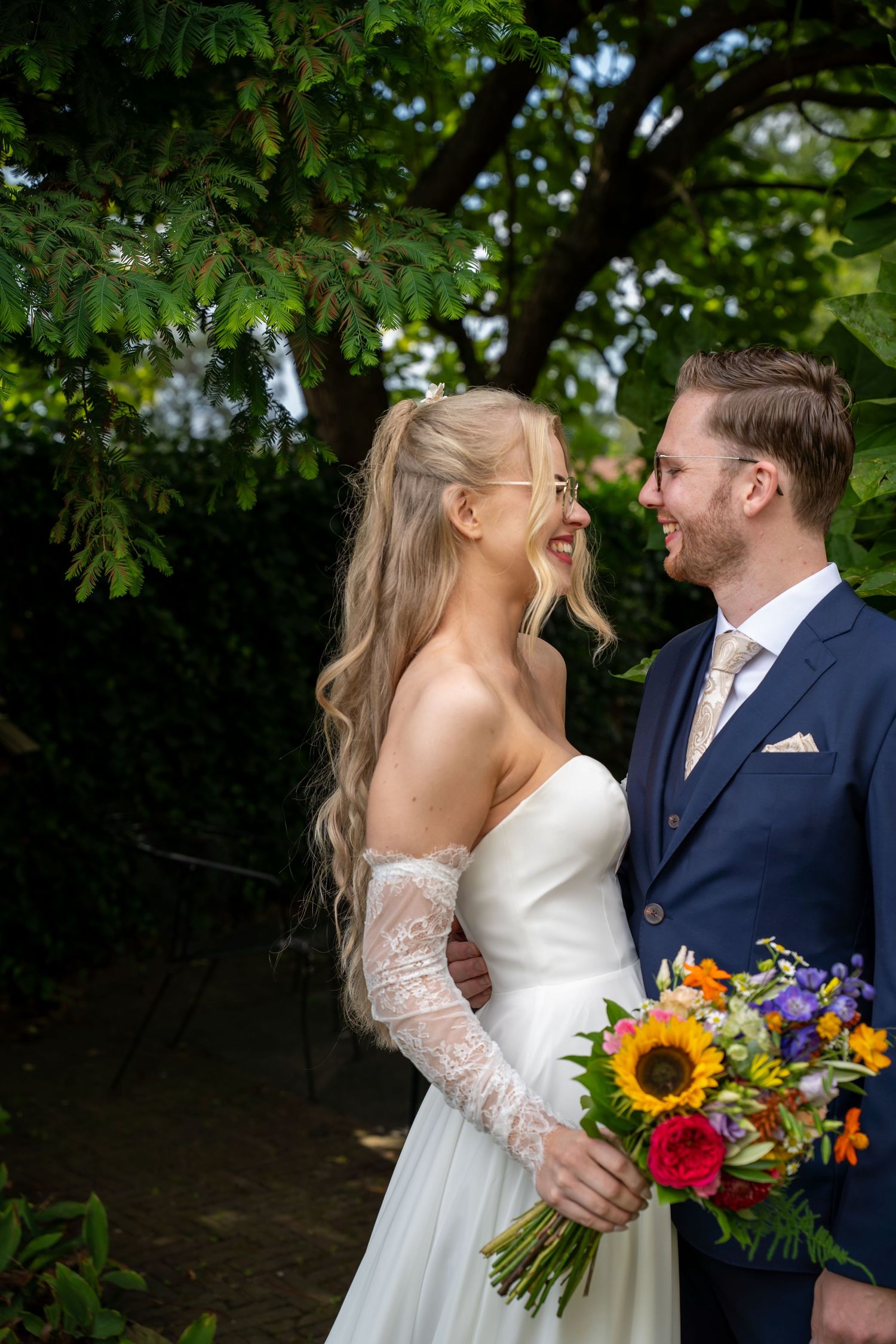 Bride and groom gazing at each other under a tree, holding a vibrant sunflower wedding bouquet.