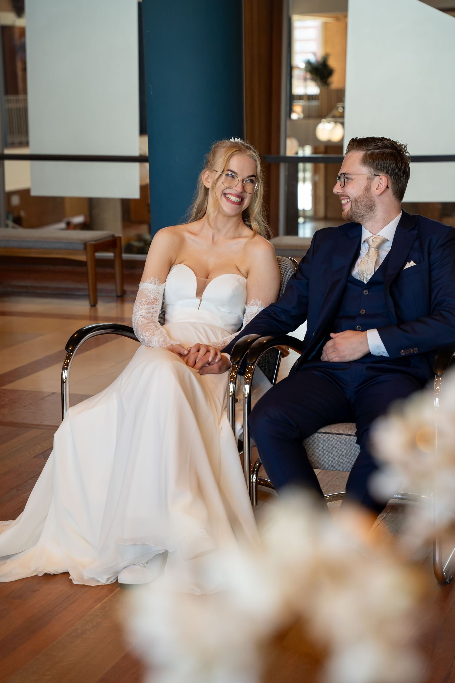 A smiling bride in a white gown and groom in navy suit sit together holding hands at their wedding reception.
