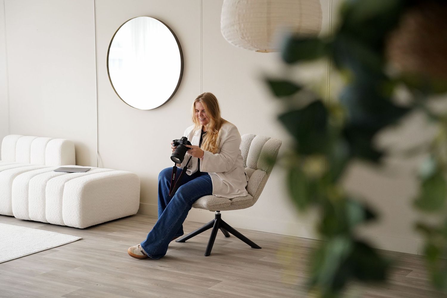 Woman sitting in a modern white chair holding a camera in a bright minimalist room with round mirror decor.