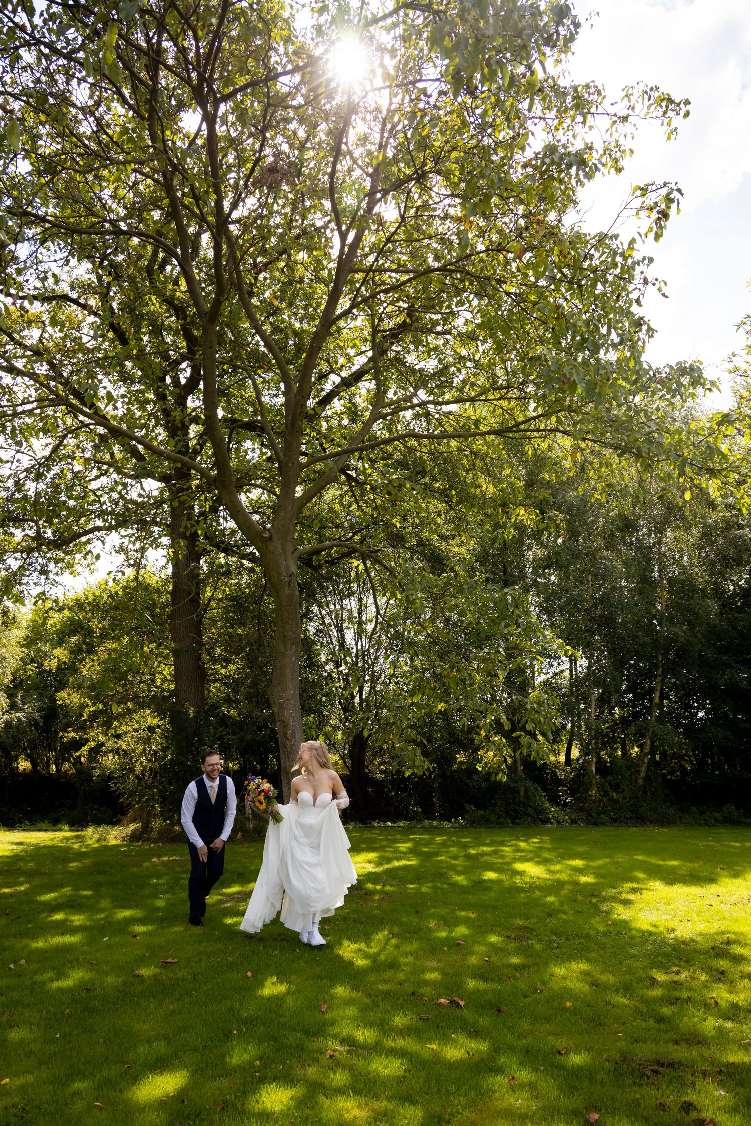 Bride and groom walking on lush green grass beneath a large sunlit tree on their wedding day.
