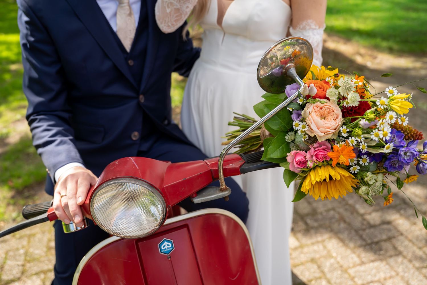 Bride and groom posing with a red Vespa scooter adorned with a colorful bouquet of sunflowers and mixed blooms.
