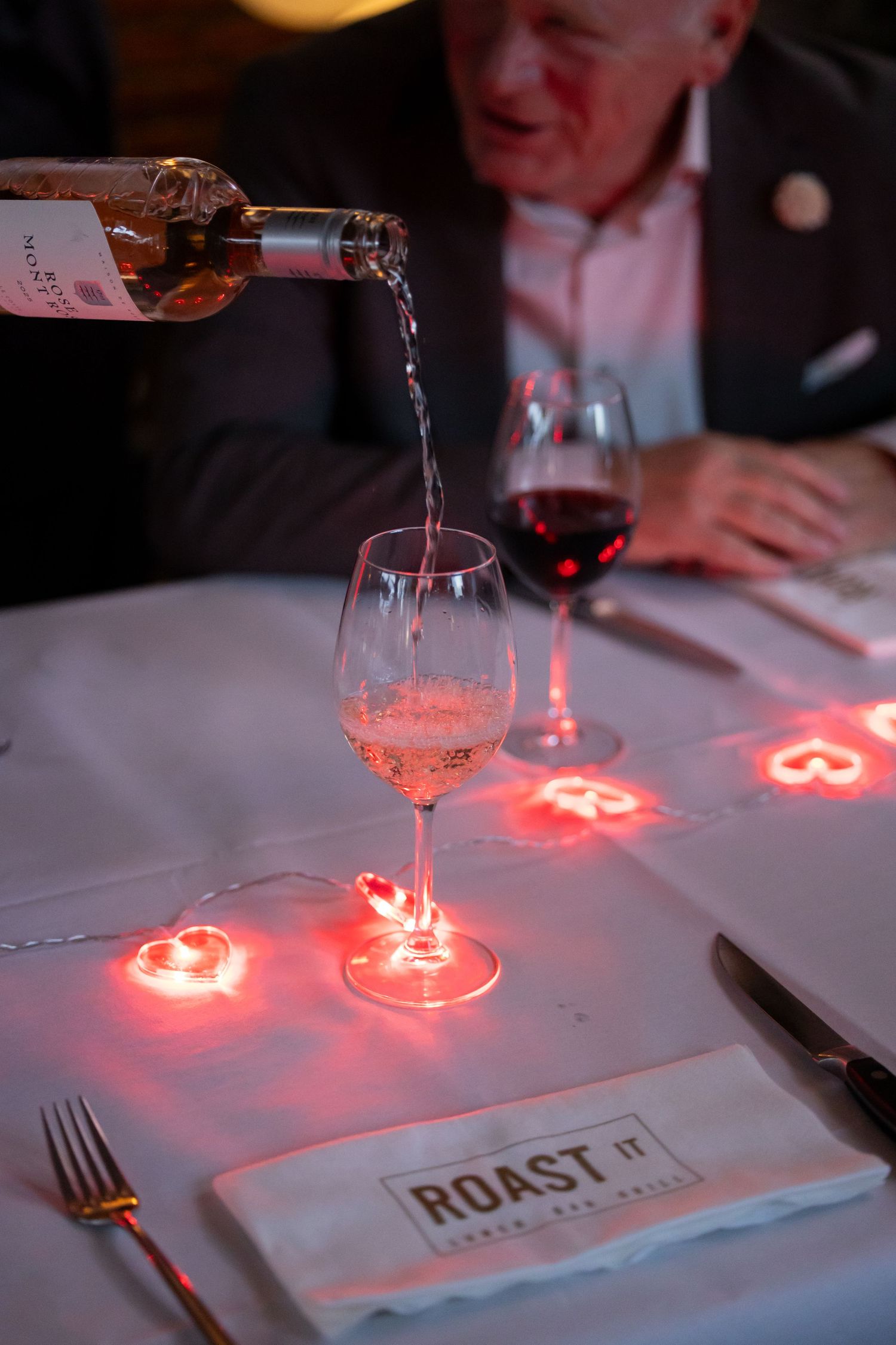 Wine being poured into a glass at a Roast It restaurant table with red heart string lights and an elderly diner.