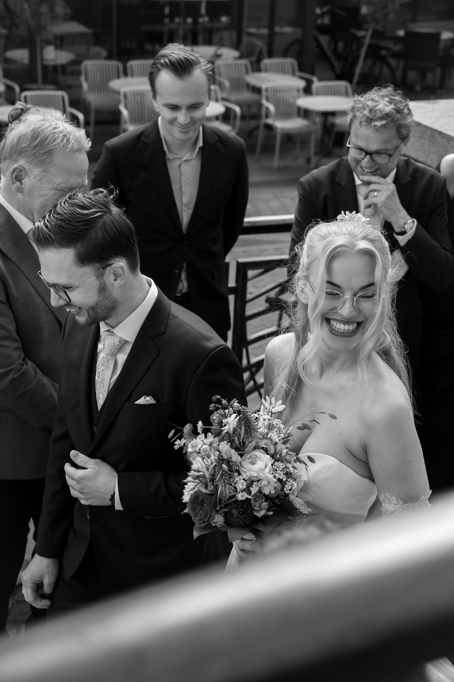Black and white photo of smiling bride holding bouquet with groom and guests on wedding day.