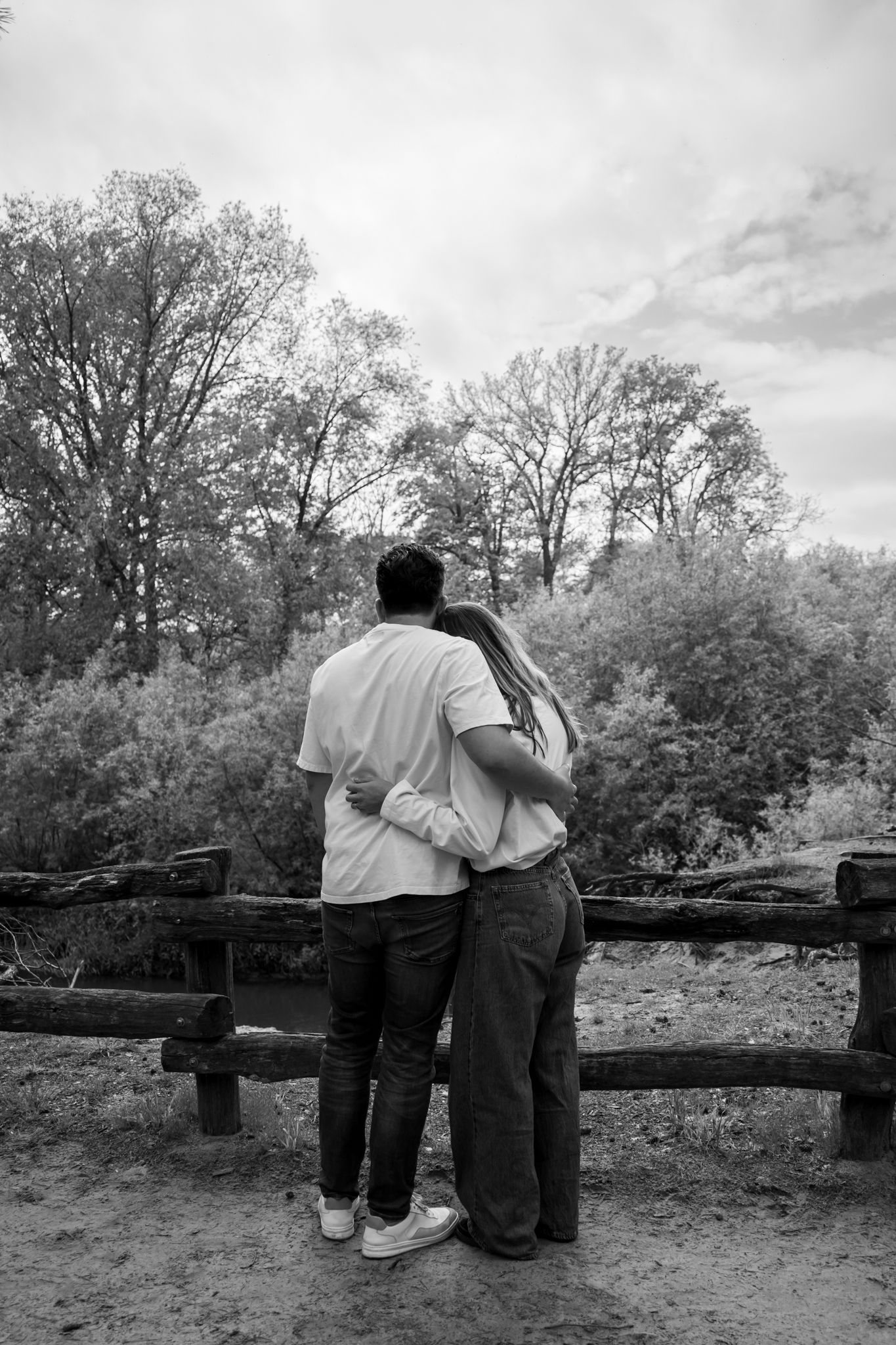 Black and white photo of a couple embracing while standing at a rustic wooden fence, gazing at lush trees.
Fleur en Timo - Koppelshoot