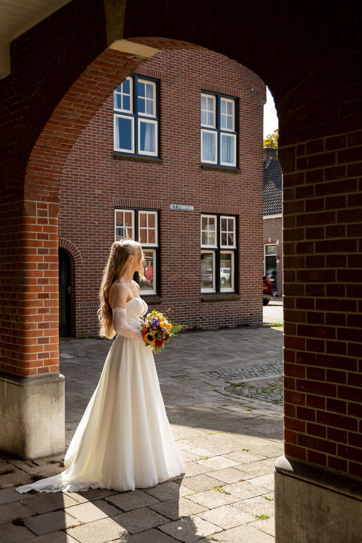 Bride in white gown holding colorful bouquet stands under brick archway in sunlit European courtyard.
