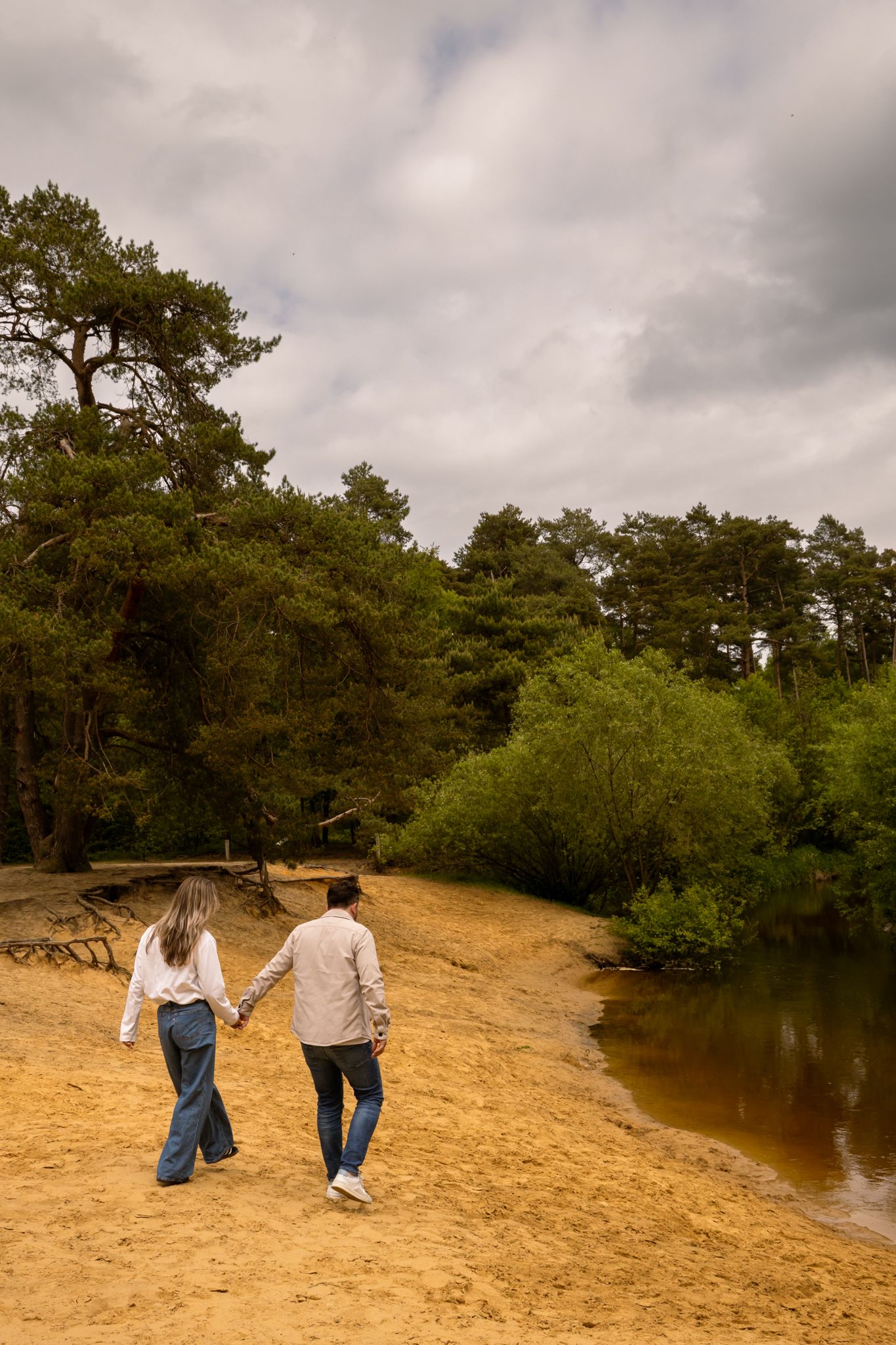 Couple holding hands walking along sandy riverbank surrounded by lush green trees under cloudy sky.
Fleur en Timo - Koppelshoot