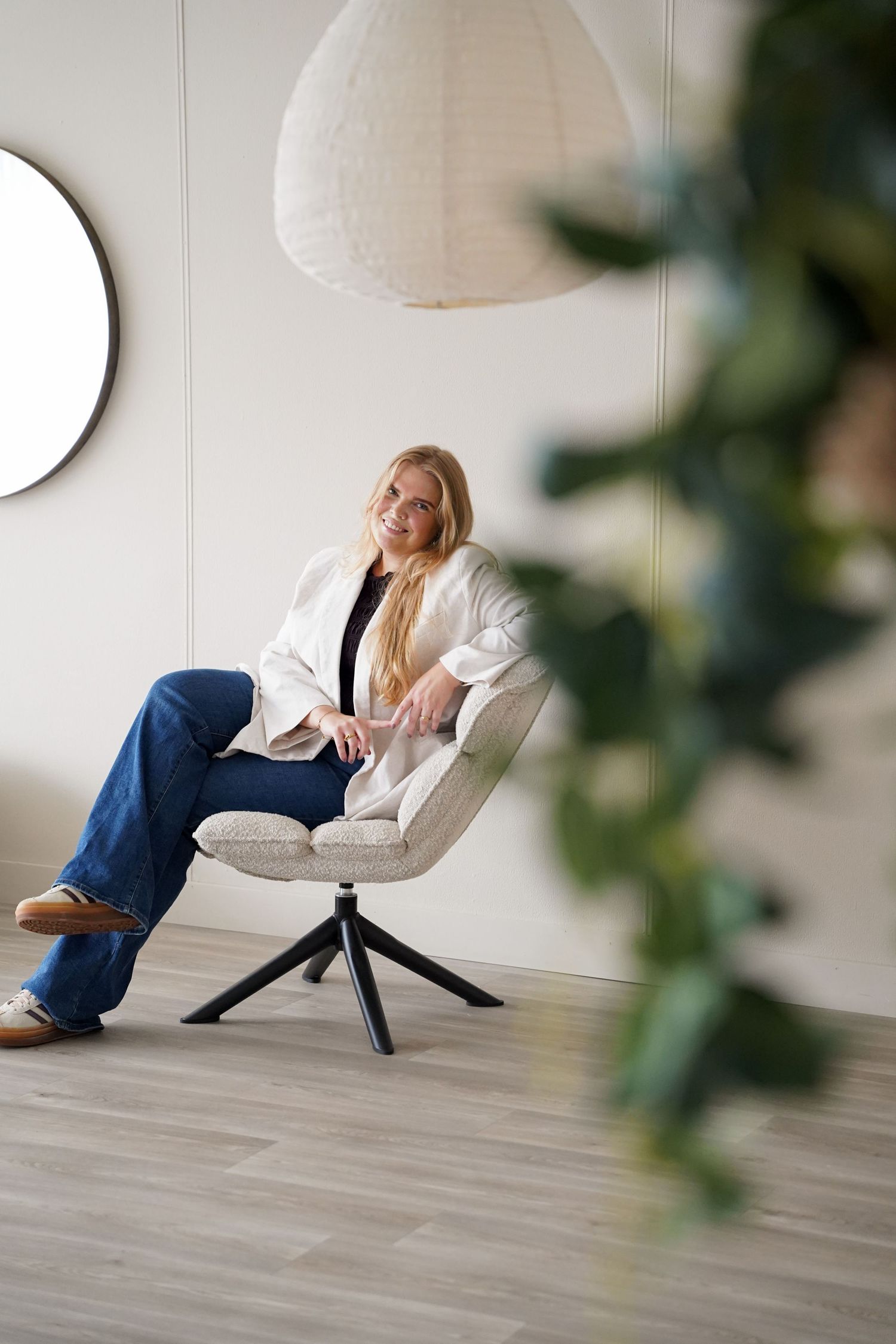 Smiling blonde woman in white blazer and jeans sitting relaxed in modern accent chair in minimalist interior.
